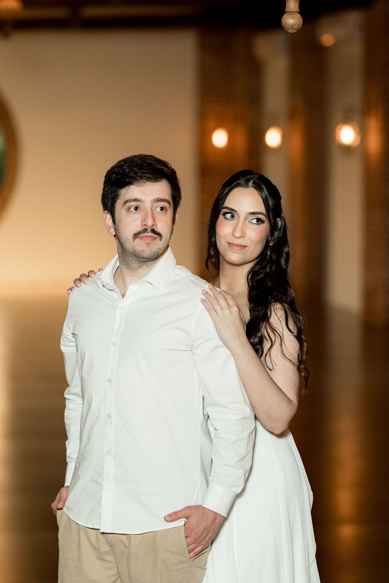 A couple in matching white outfits pose elegantly in a warmly lit venue, the woman resting her hand on the mans shoulder.