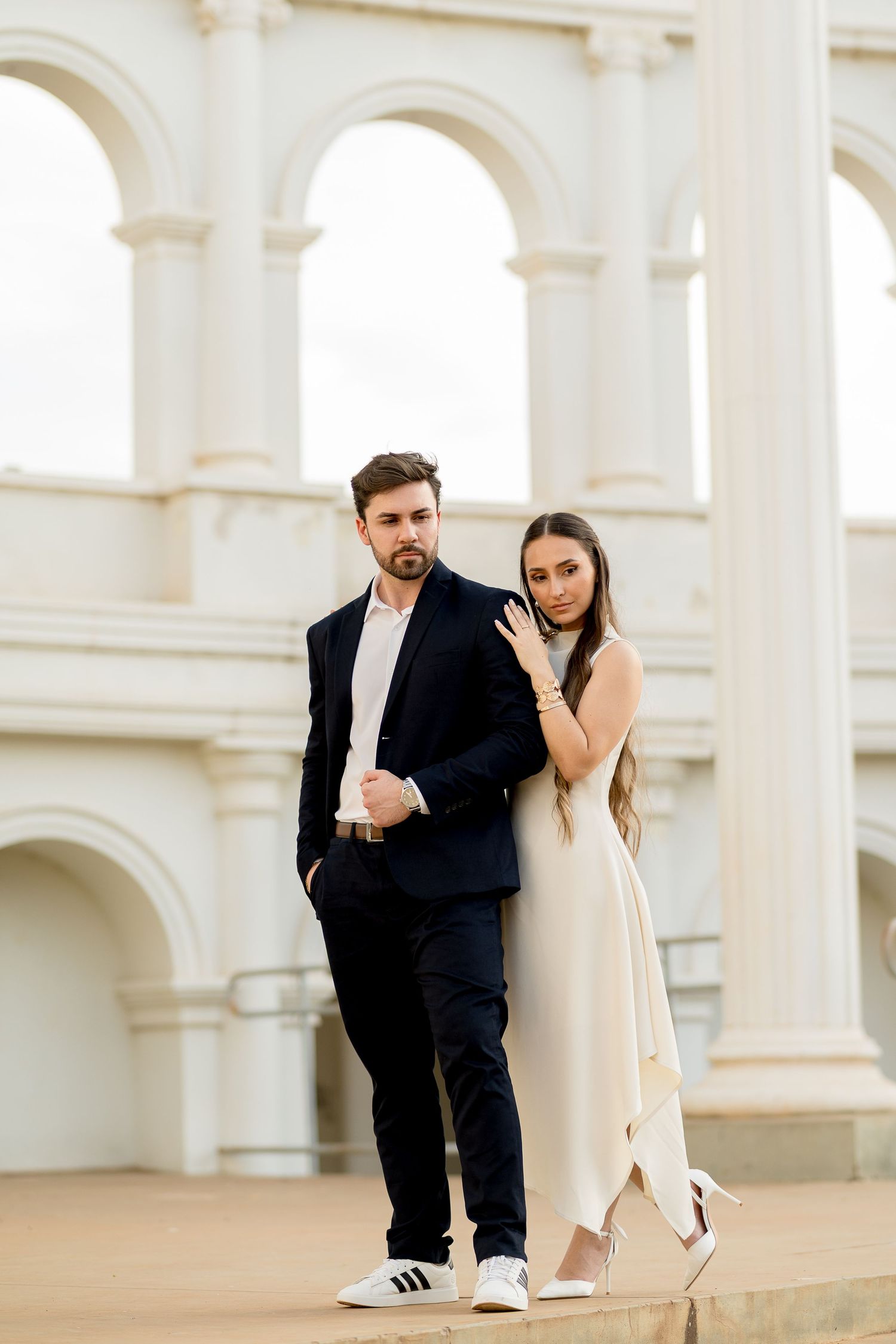 Elegant couple posing before white arched building, man in dark suit, woman in cream dress and heels.