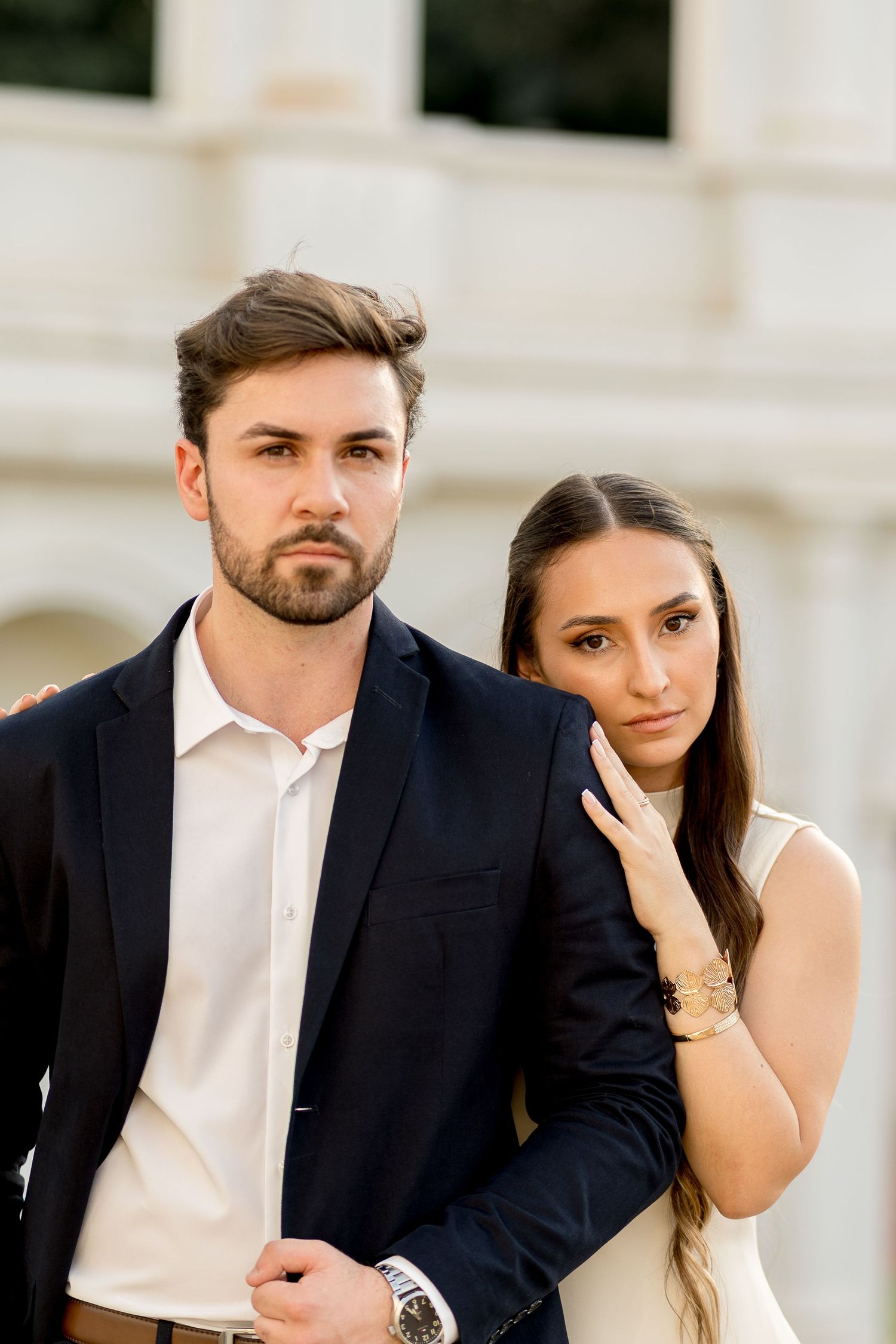 Elegant couple in formal attire posing outdoors, man in dark suit with white shirt, woman in white dress leaning on his shoulder.