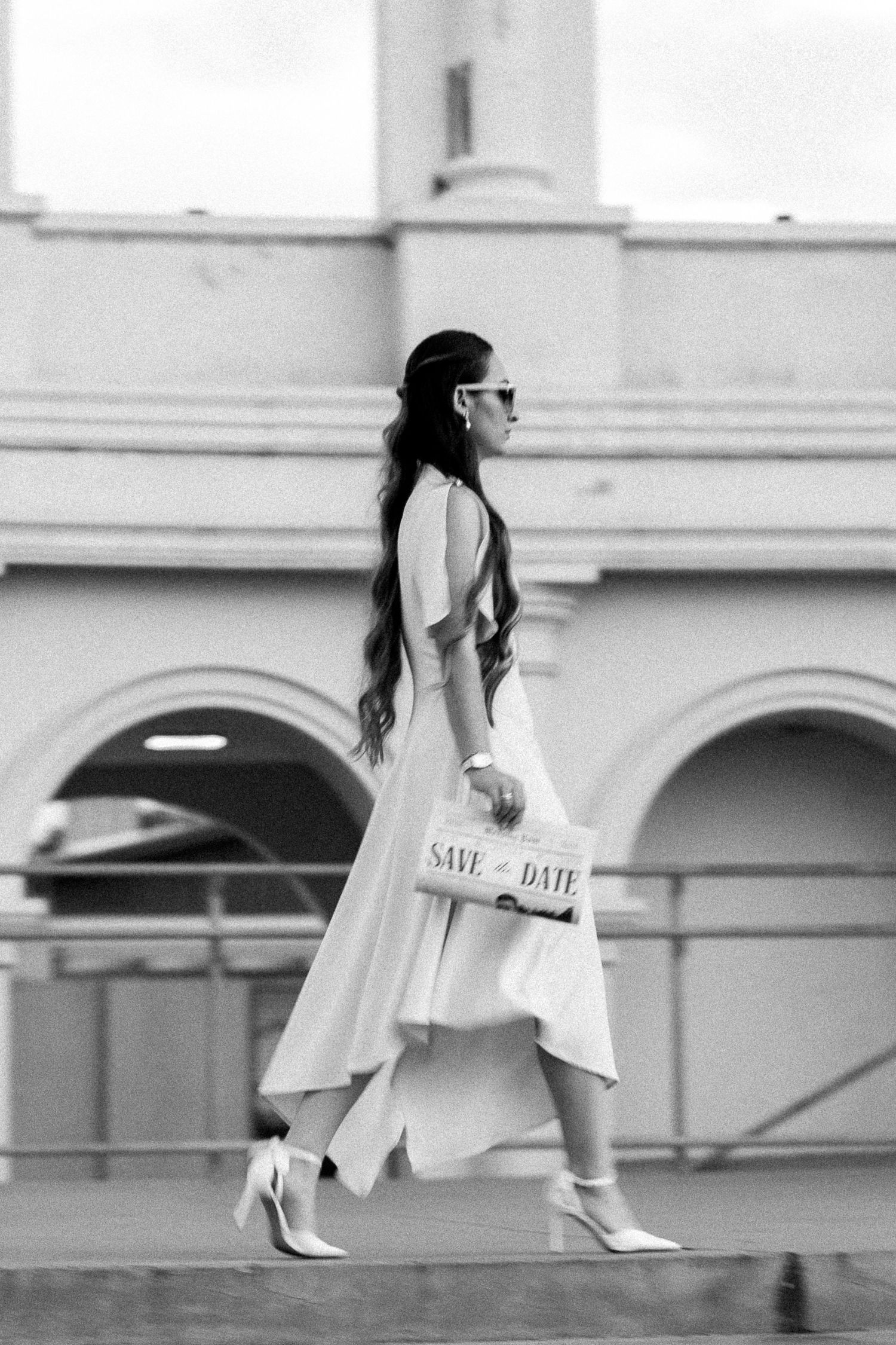 Black and white photo of elegant woman in white dress holding Save the Date clutch, walking past arched building.