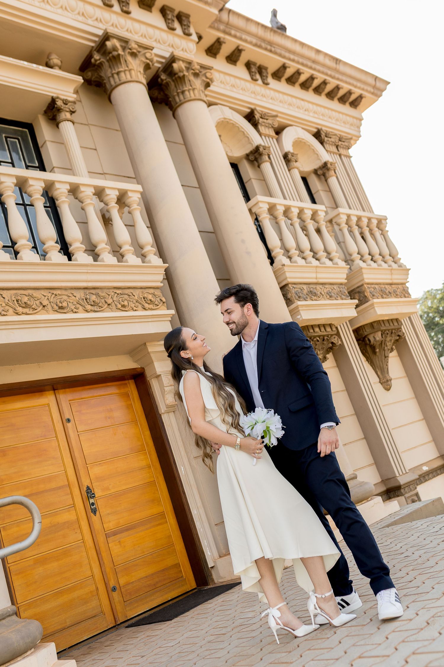 Bride and groom pose in front of a grand neoclassical building with tall columns and ornate balustrades.
