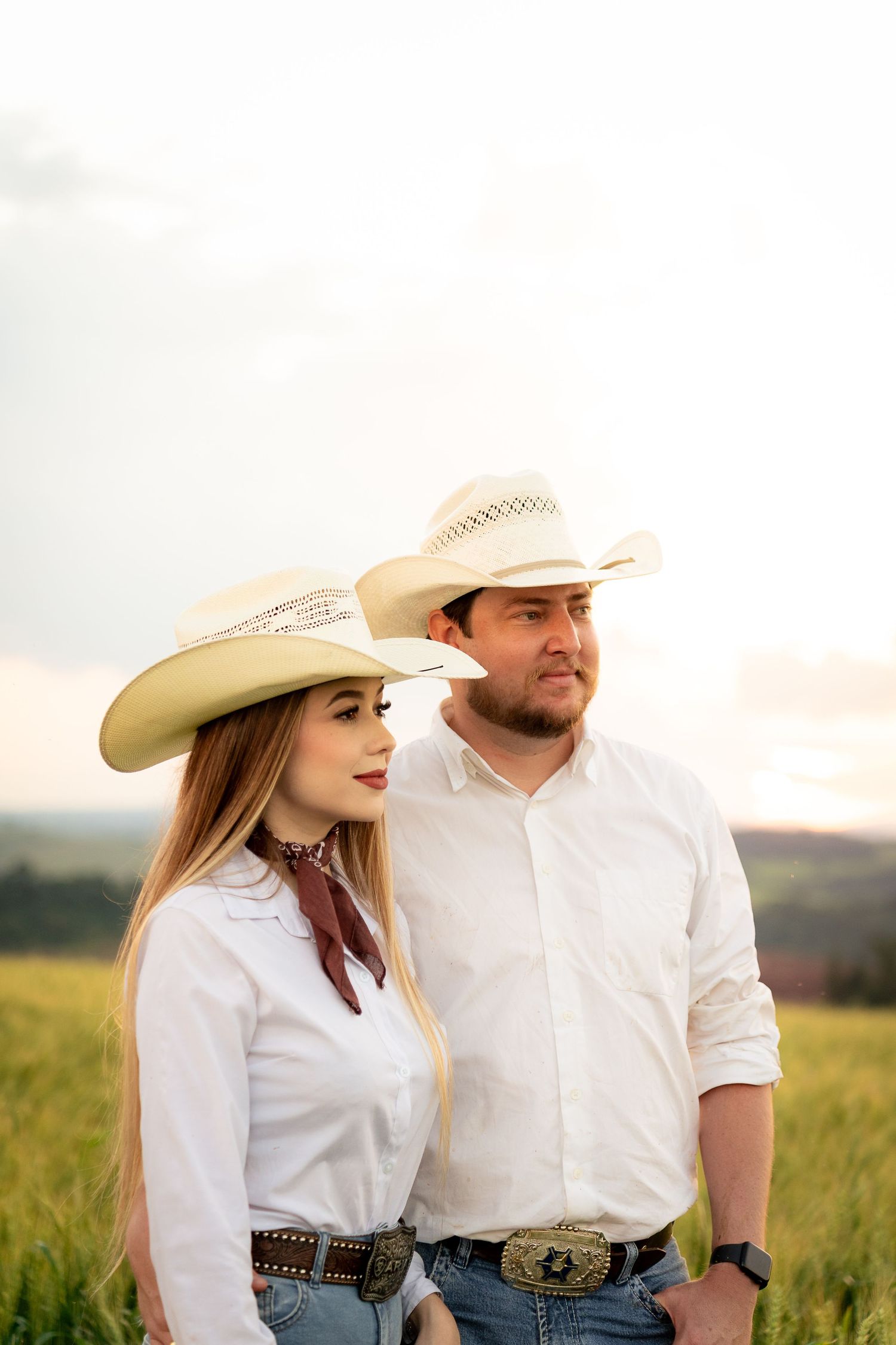A couple in matching white shirts and cowboy hats stand together in a green field at sunset.