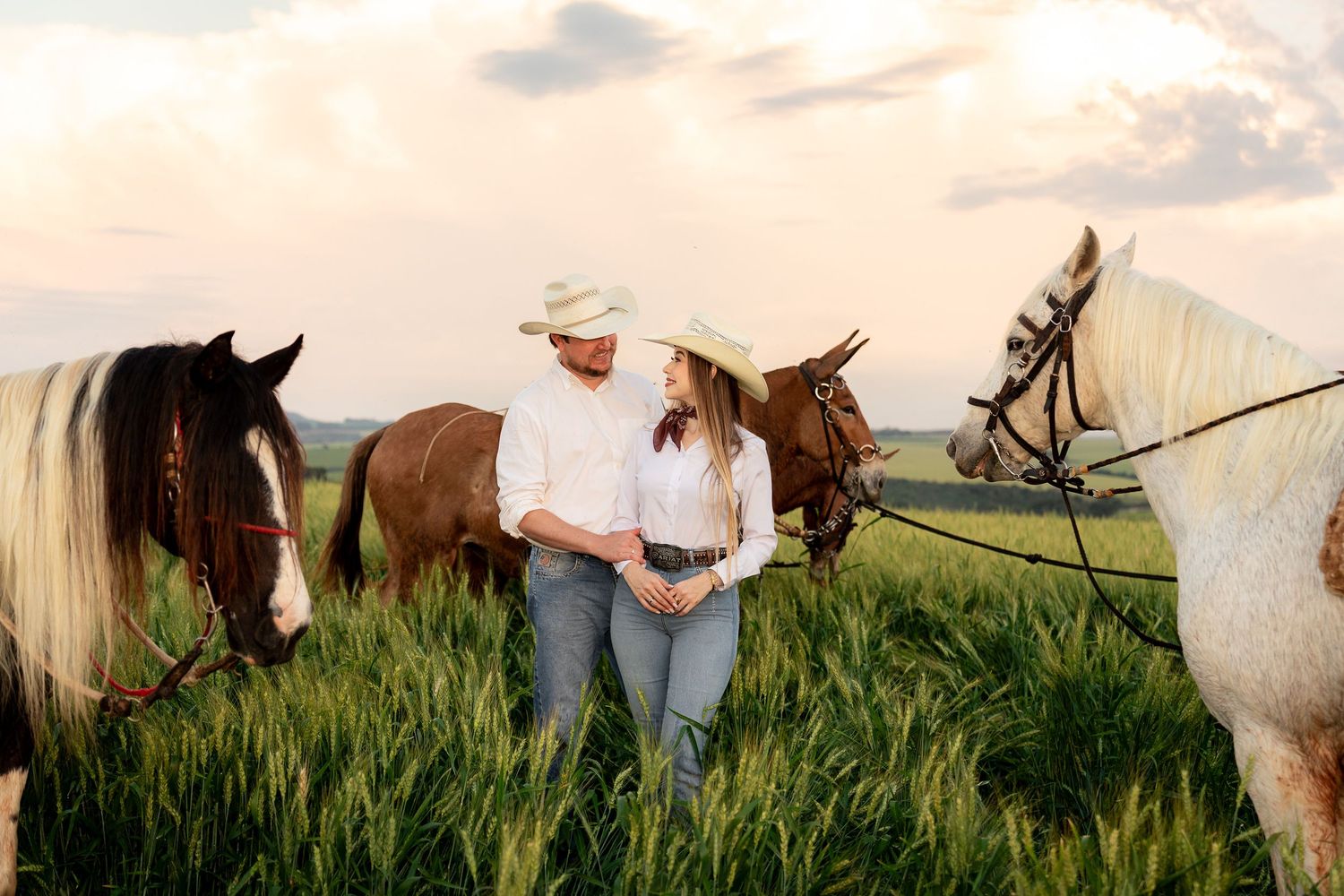 Couple in cowboy hats standing among horses in green field at sunset.
