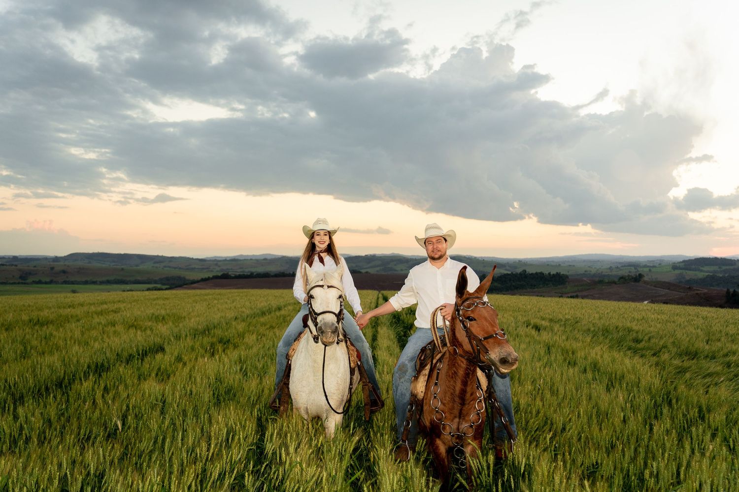 Couple in cowboy hats riding horses through a lush green field at sunset with dramatic cloudy sky.