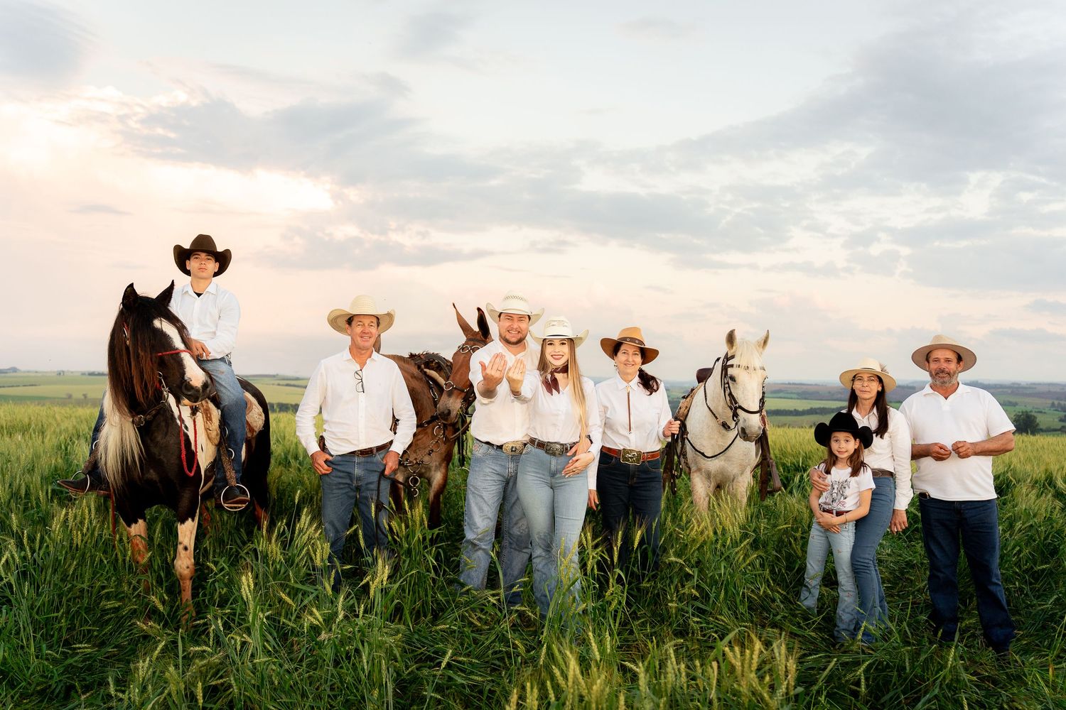 Family in cowboy hats posing with horses in a green field at sunset, wearing western attire for a country-style portrait.