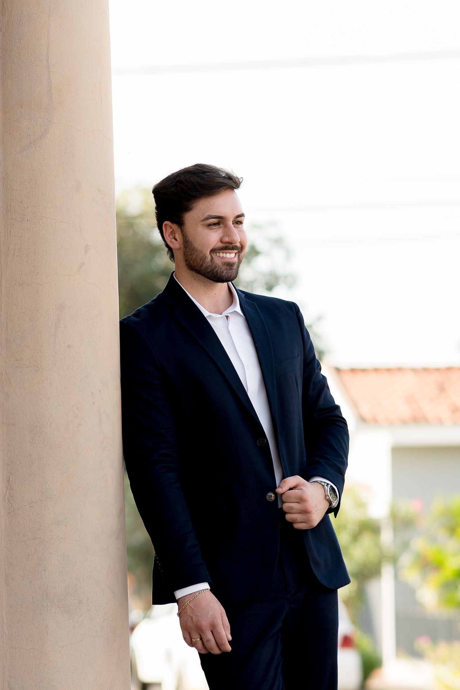 Smiling young man in navy suit leaning against column outdoors.