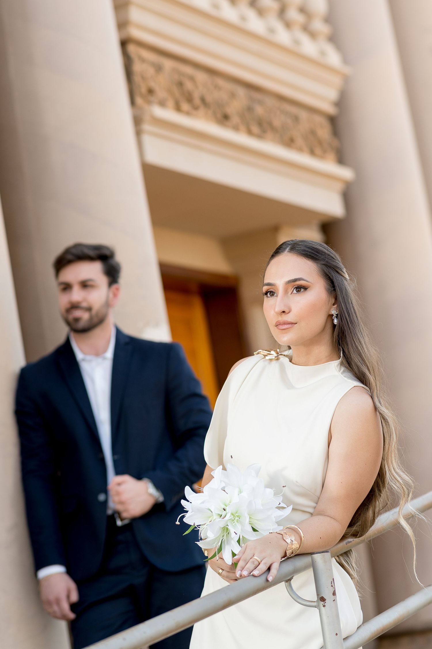 Bride in white dress holding lily bouquet stands by classical columns, groom in dark suit behind her.