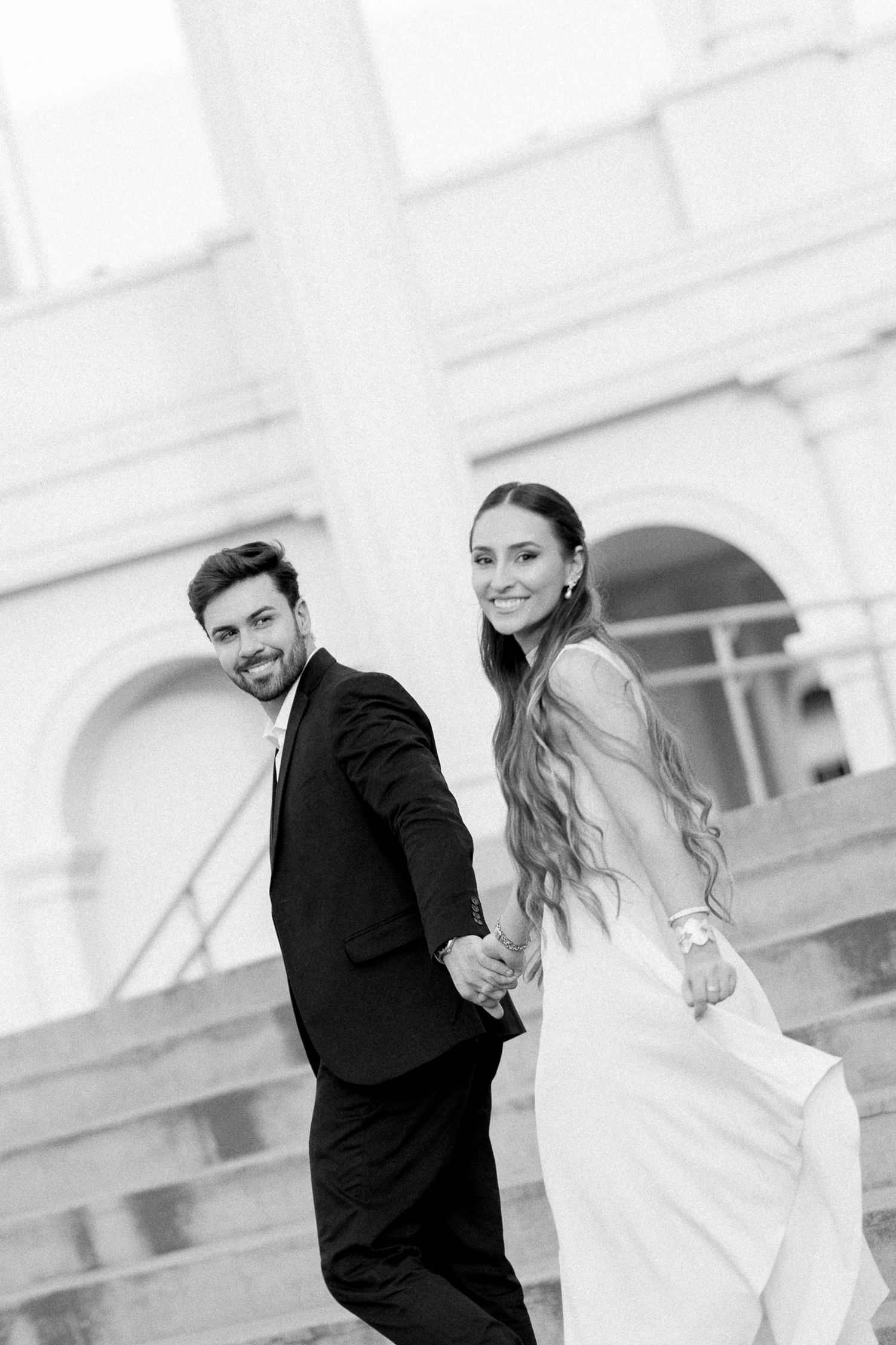 Black and white photo of a smiling bride and groom holding hands on steps outside a grand building.