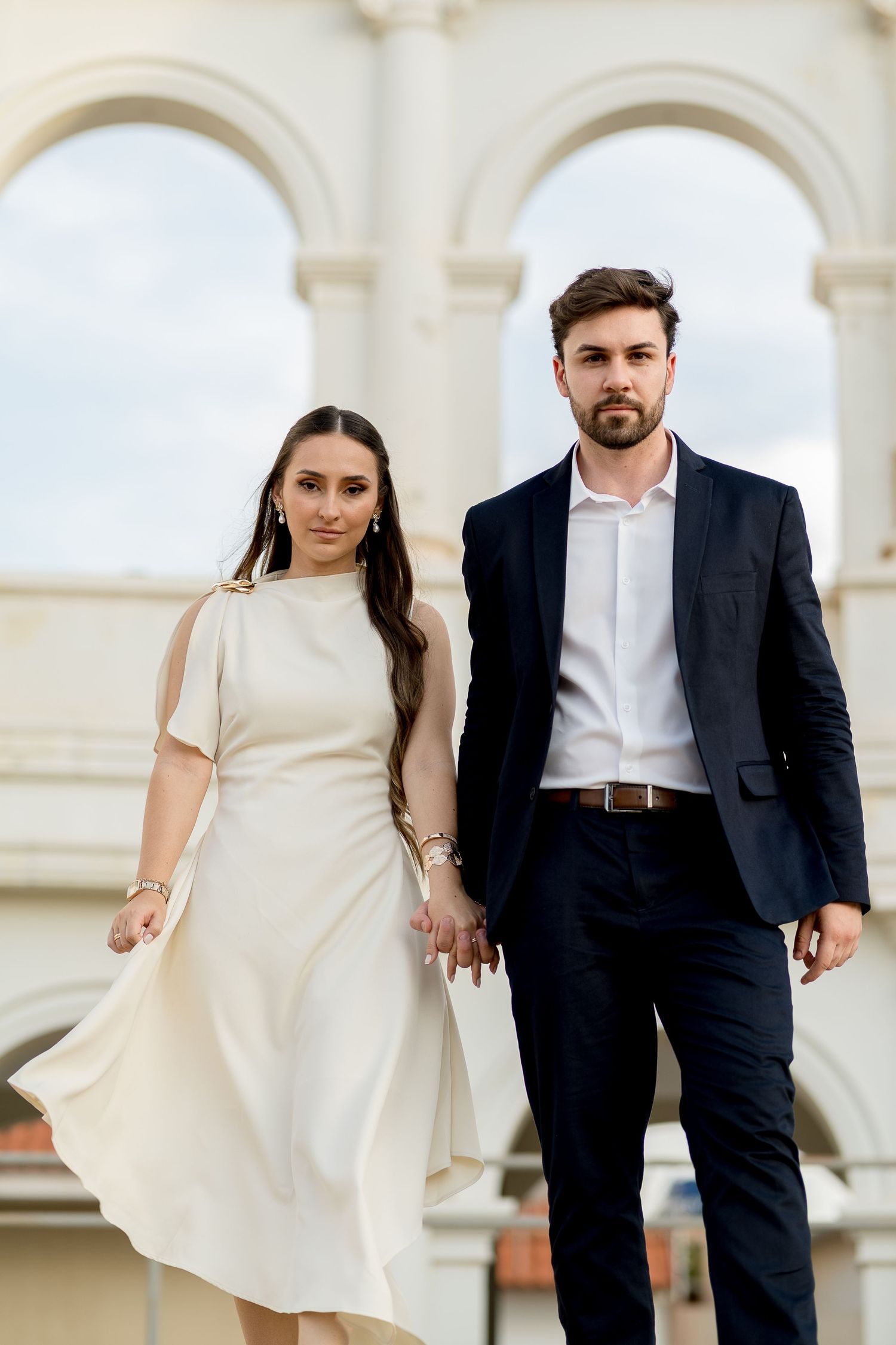Elegant couple holding hands, woman in white dress, man in dark suit, posing before grand arched architecture.