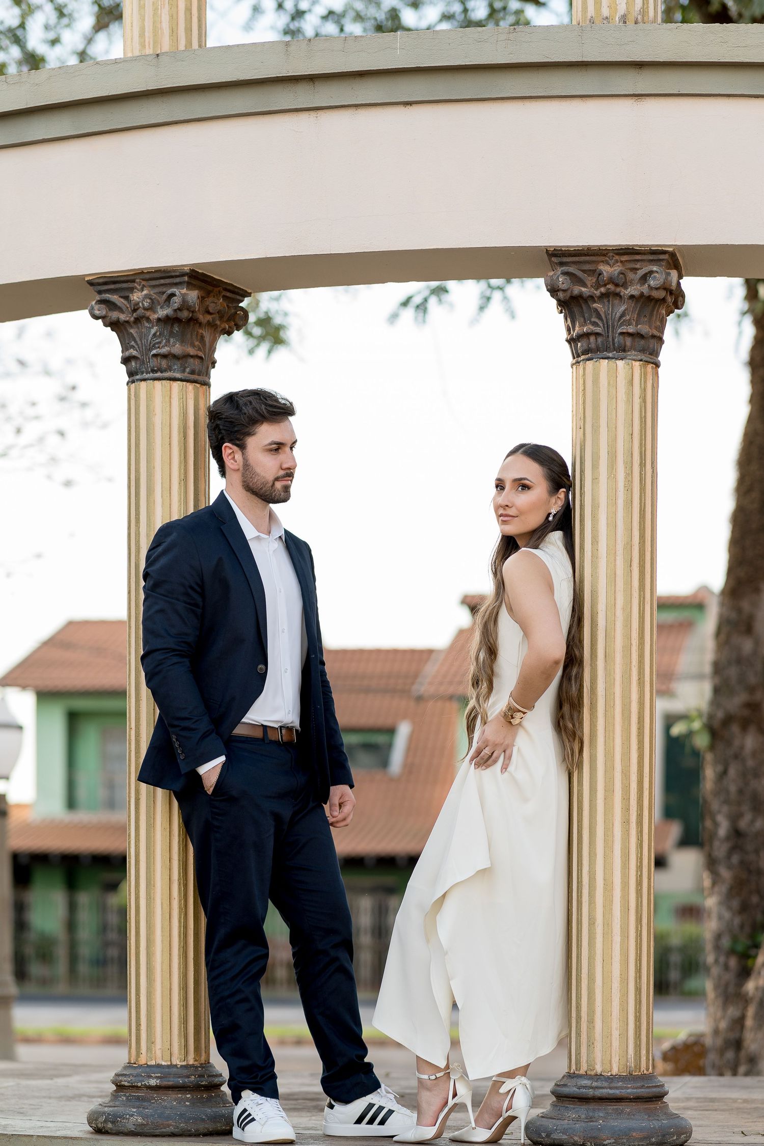 Stylish couple standing between ornate golden columns, man in navy suit, woman in white backless dress with heels.
