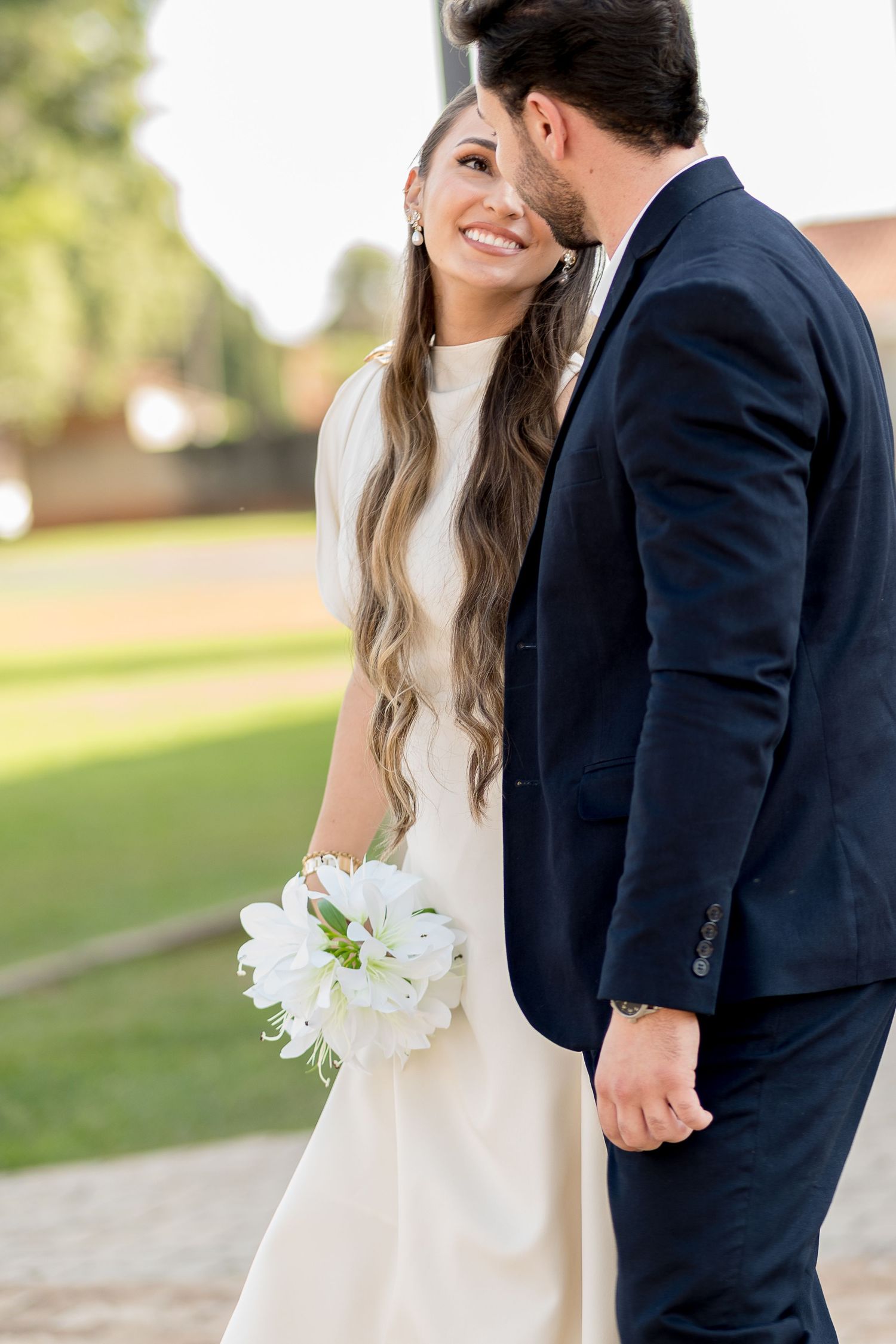 Smiling bride in white dress holding white flowers stands with groom in navy suit outdoors.