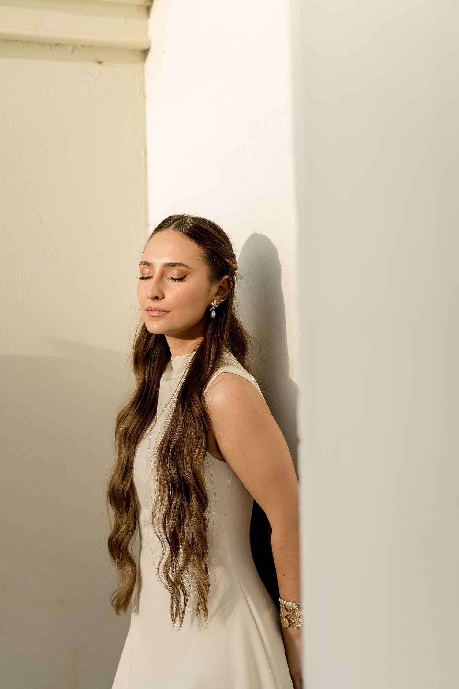 Young woman with long wavy hair in white dress leaning against white wall with eyes closed.