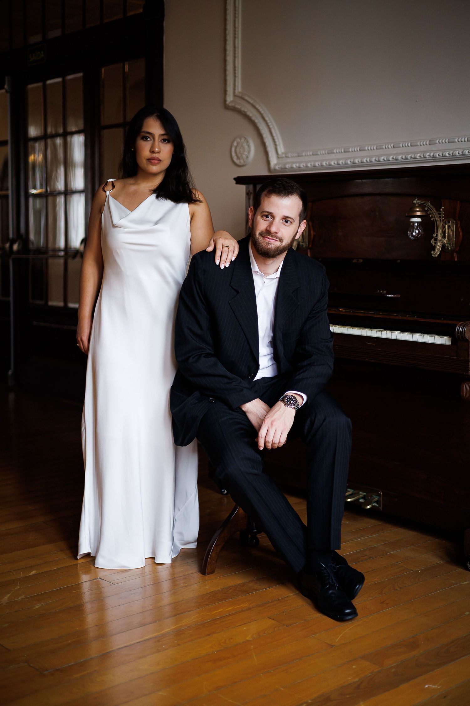 Elegant couple posing by a grand piano, woman in white gown, man in black suit, in a classic interior.