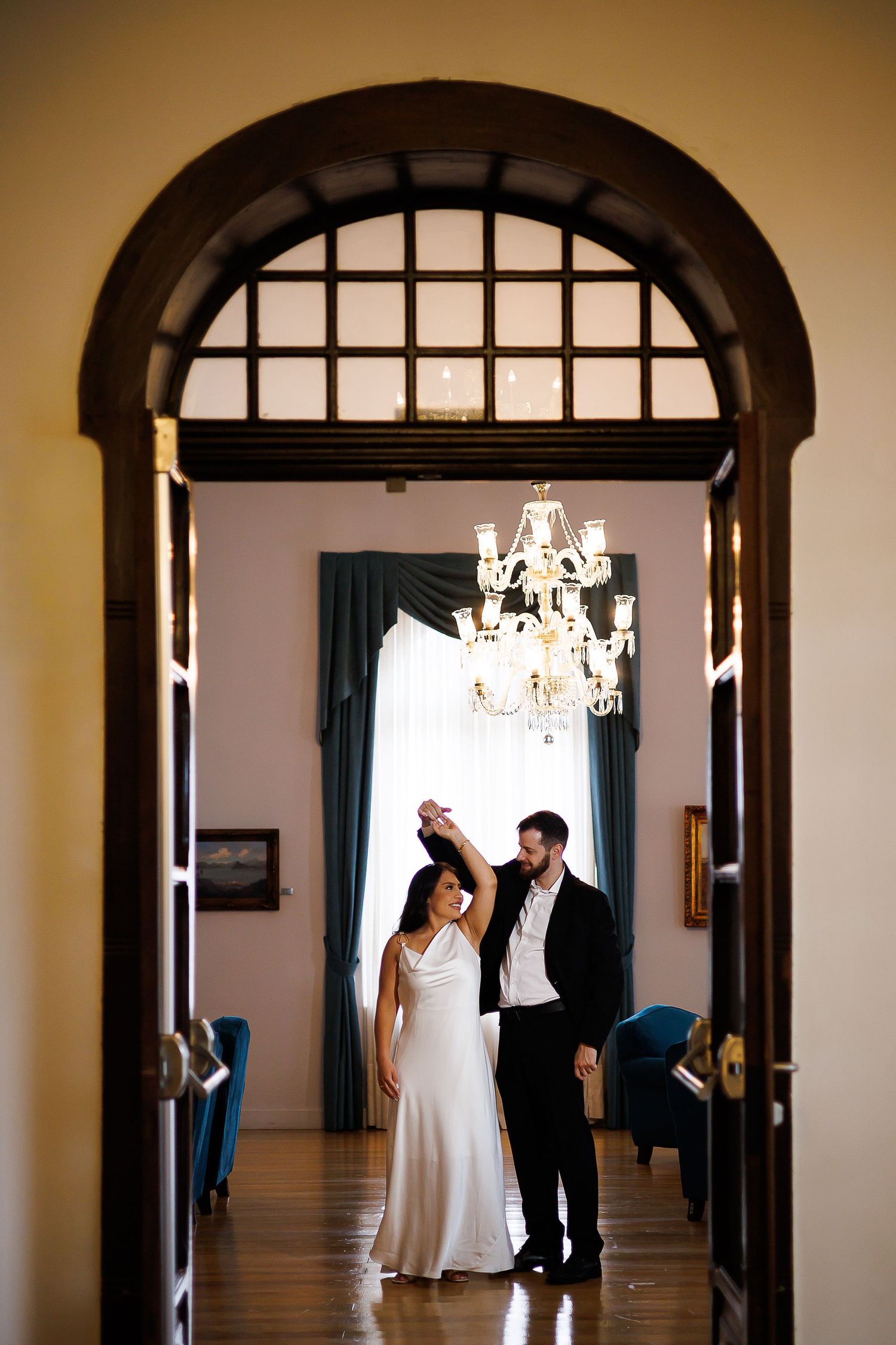 Bride and groom dancing elegantly under a crystal chandelier in a grand archway doorway.