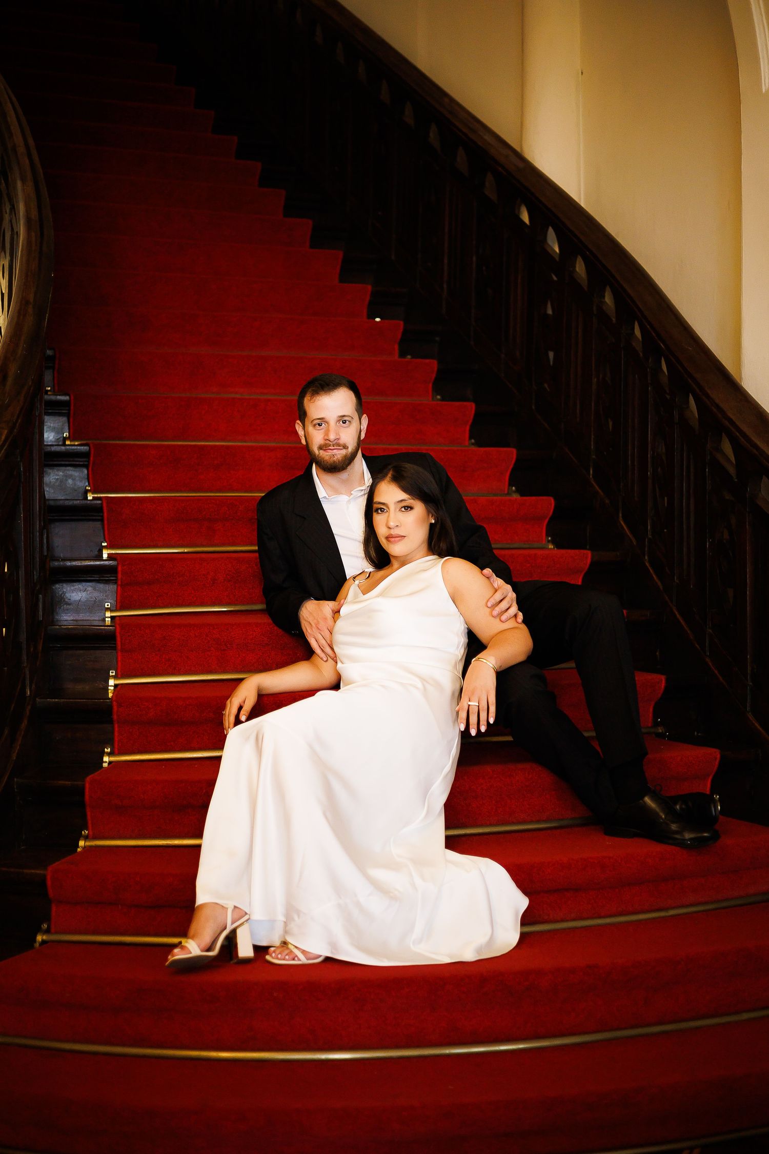 Couple in formal attire posing on red carpeted staircase, woman in white gown seated with man in black suit behind her.