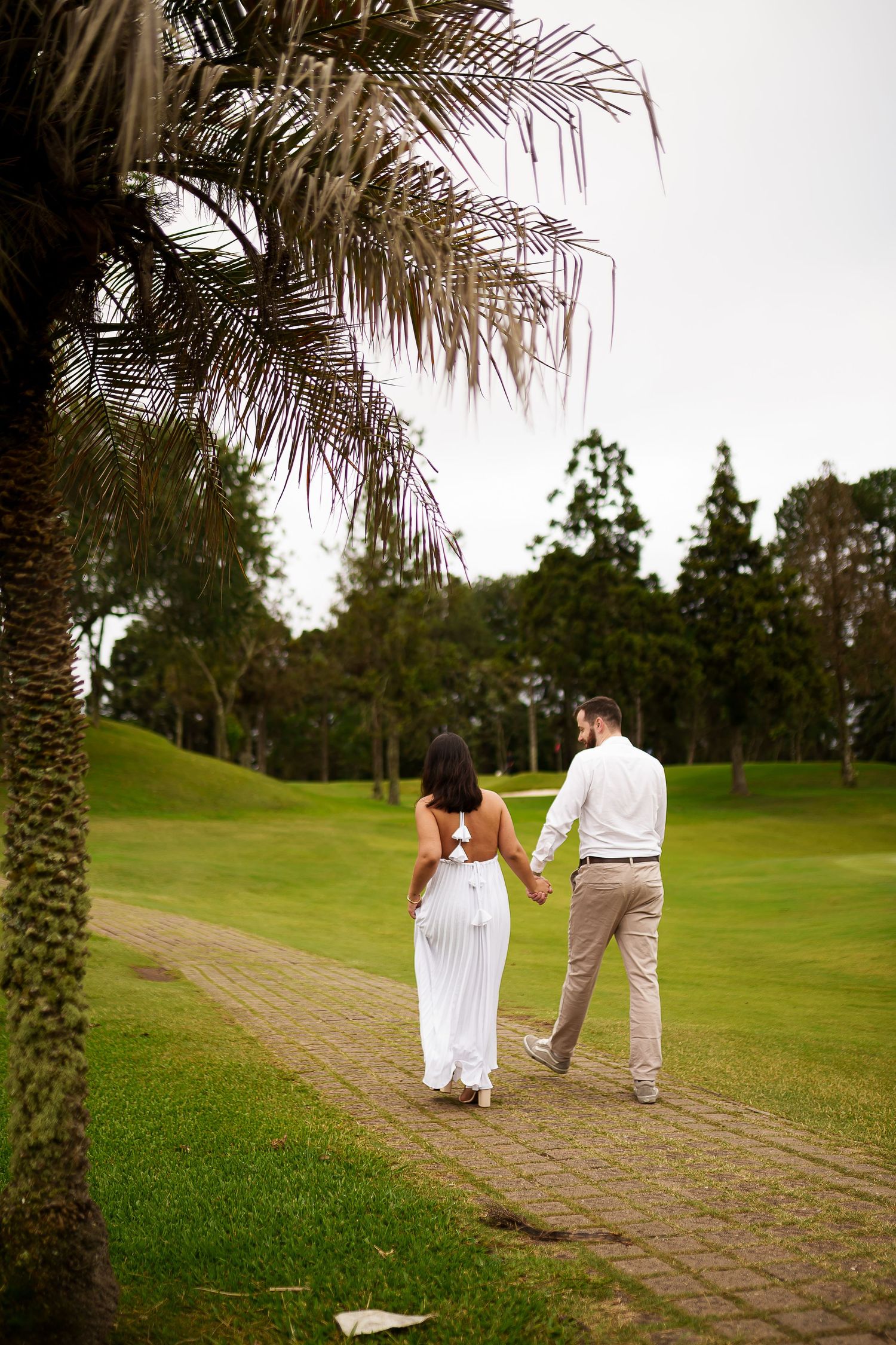 Couple holding hands walking on a brick path through lush green park, woman in white dress, man in white shirt.