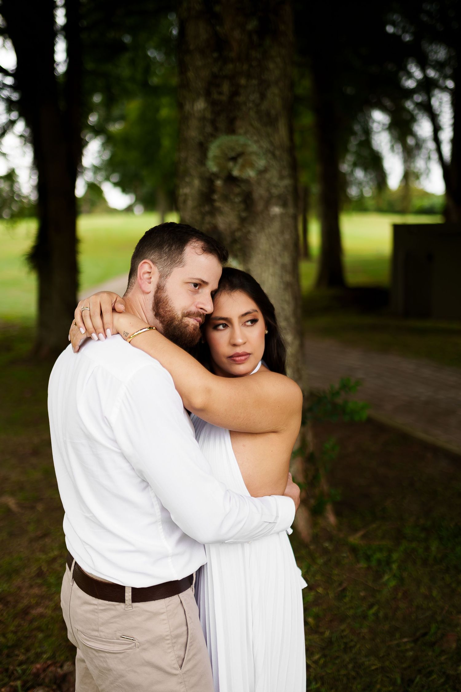 A couple in white attire embraces tenderly outdoors, surrounded by lush green trees in a romantic setting.
