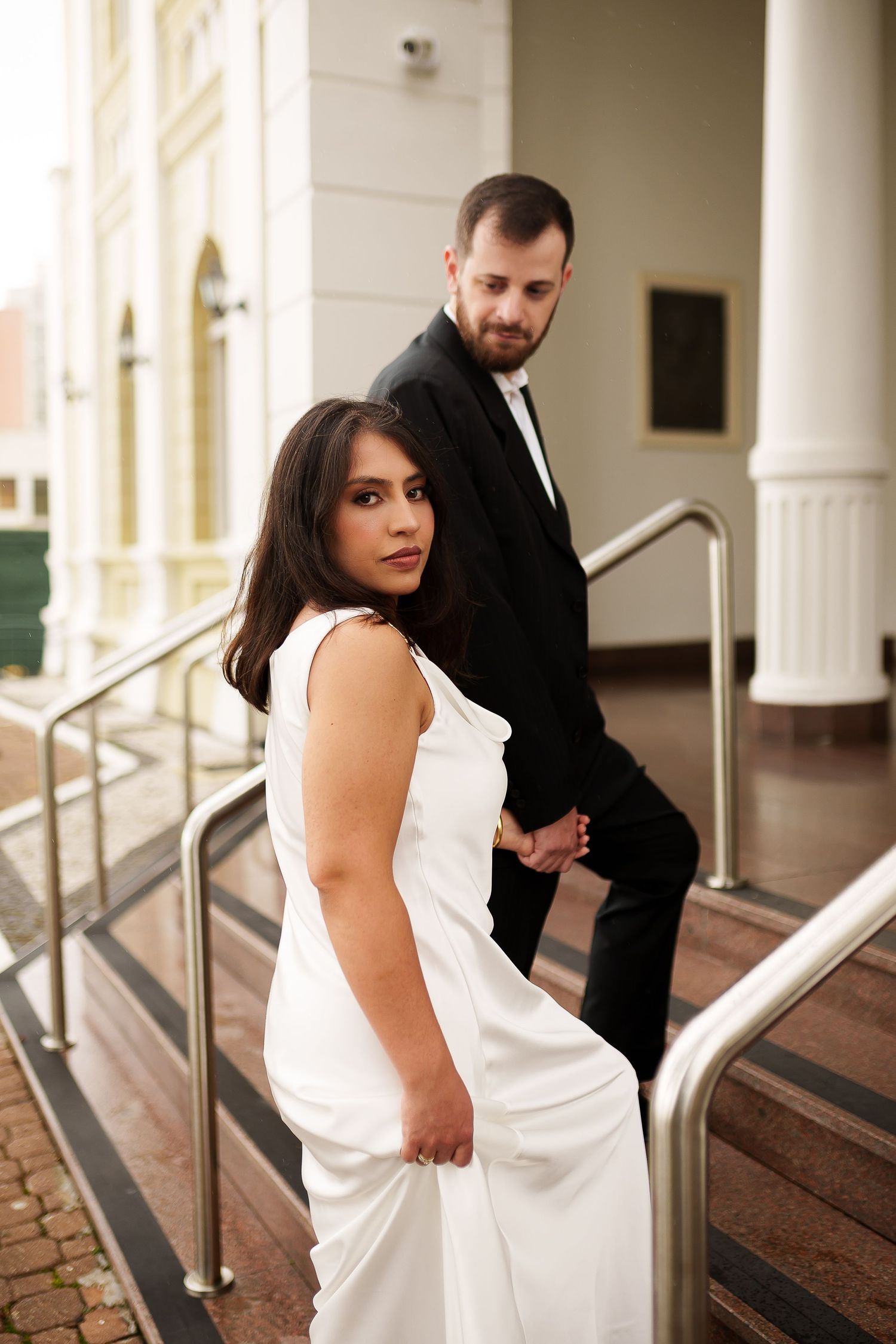 Elegant couple in formal attire on steps, bride in white gown holding hands with groom in black suit.