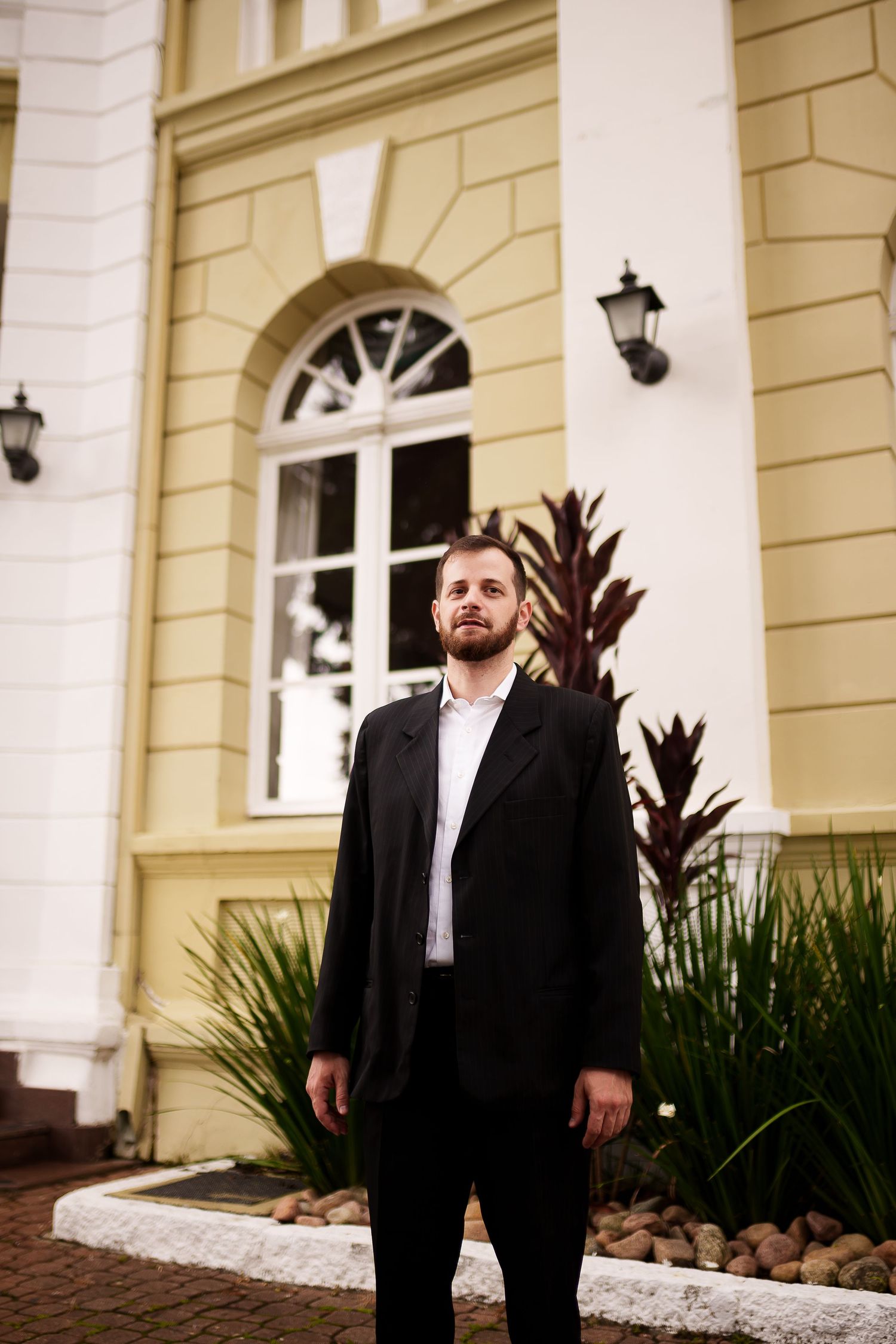 Bearded man in black suit standing confidently outside elegant yellow and white building with arched window.