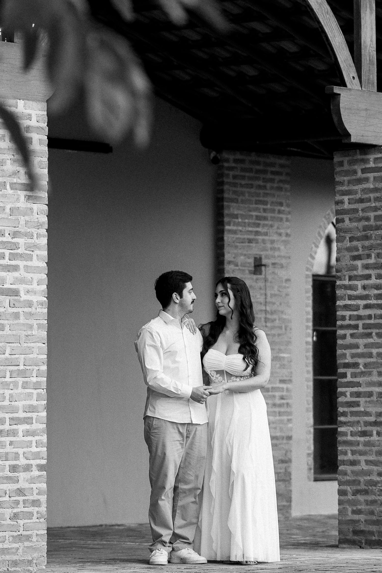 Black and white photo of a couple holding hands and gazing at each other near a brick building.
