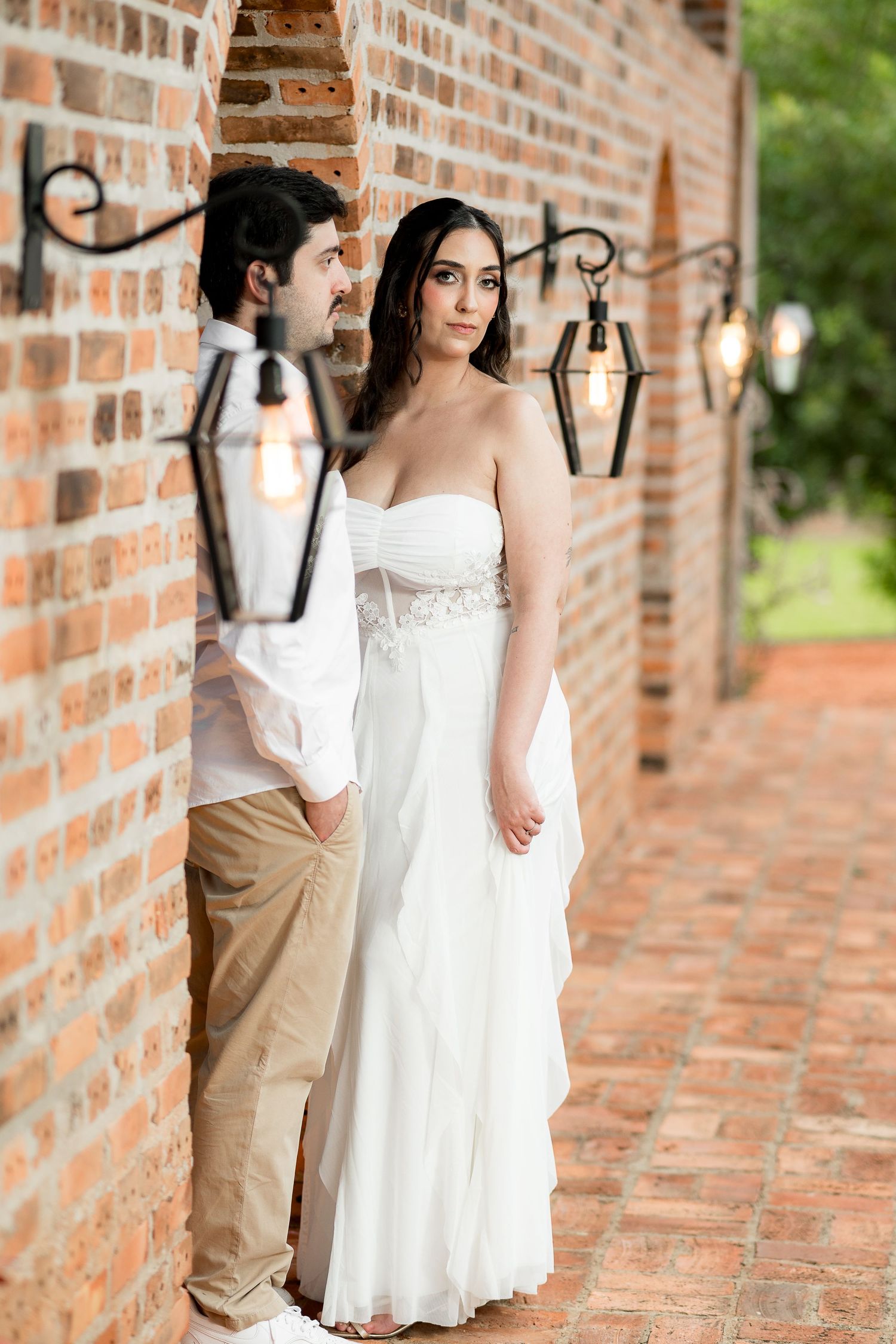 Bride in strapless white gown and groom in casual attire lean against brick wall with vintage lanterns.