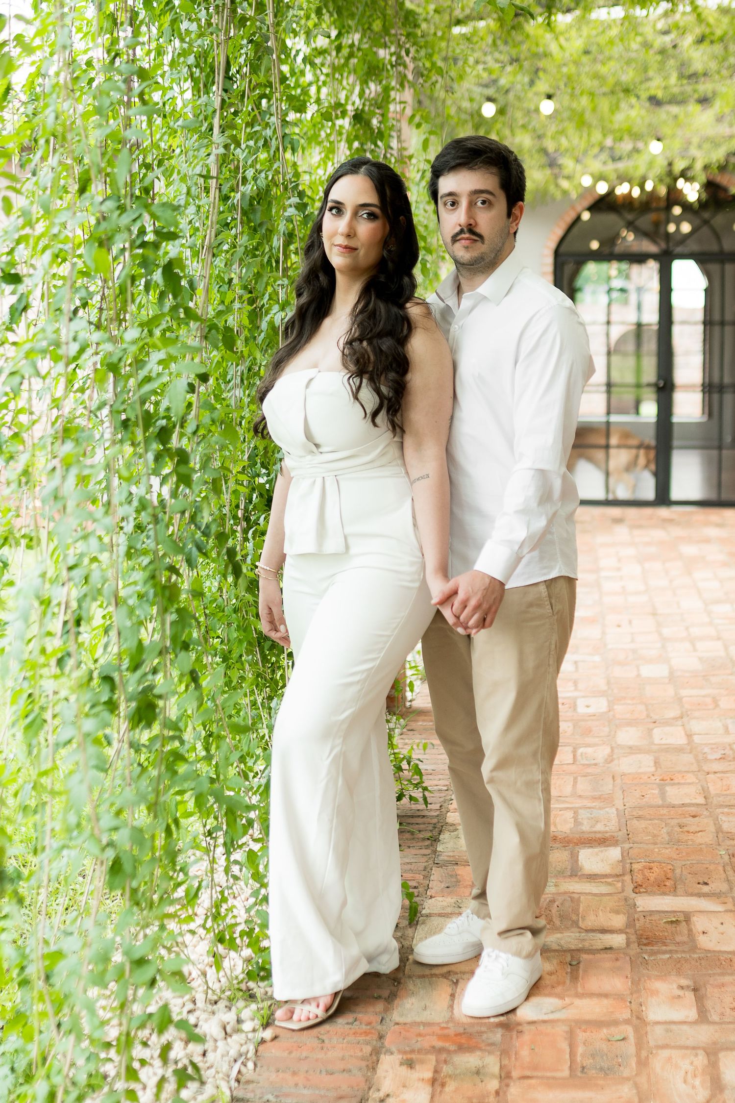 Couple in white outfits posing together in a lush garden with brick pathway and hanging greenery.