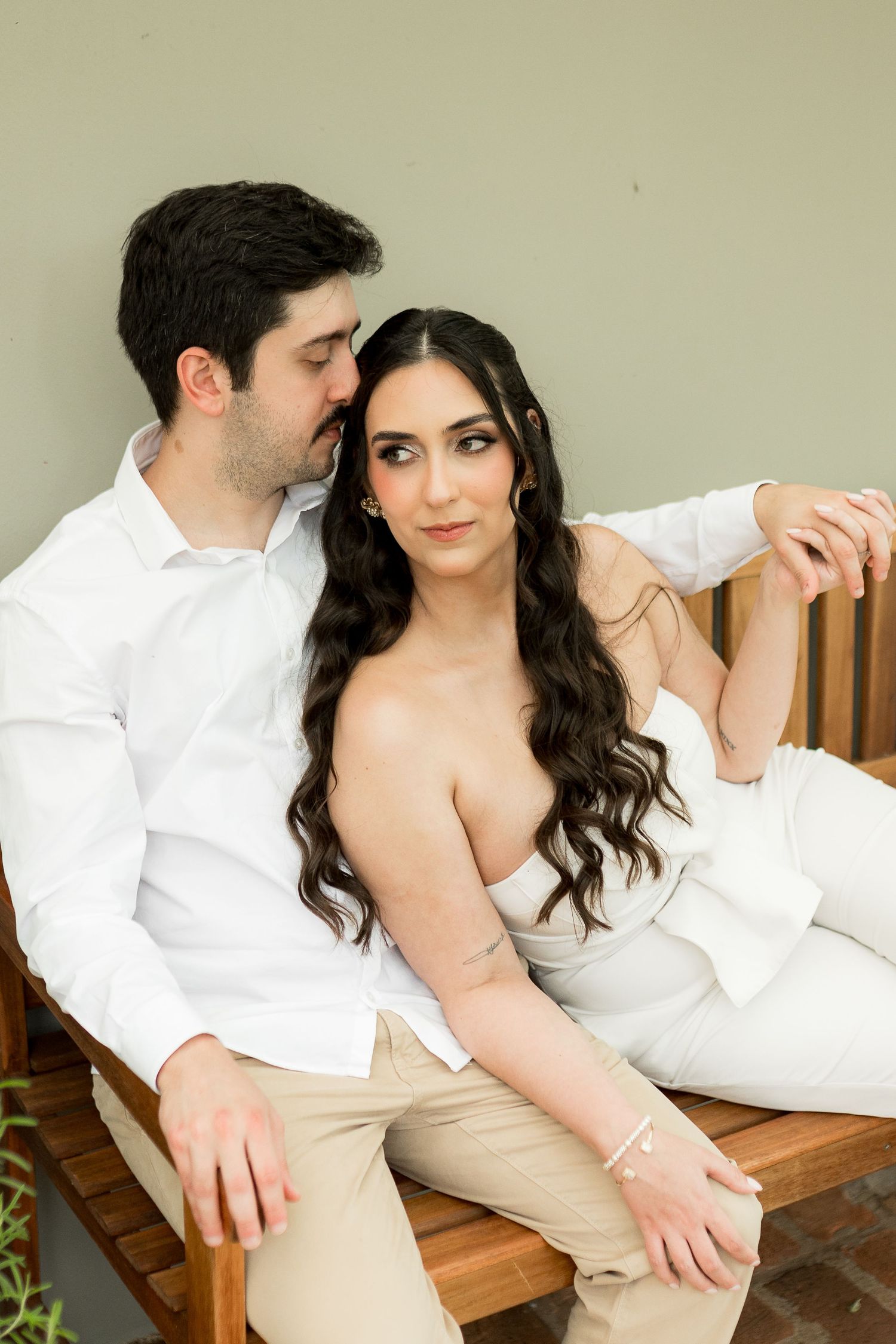 A couple sits on a wooden bench, man in white shirt kissing woman's cheek, she wears a white outfit.