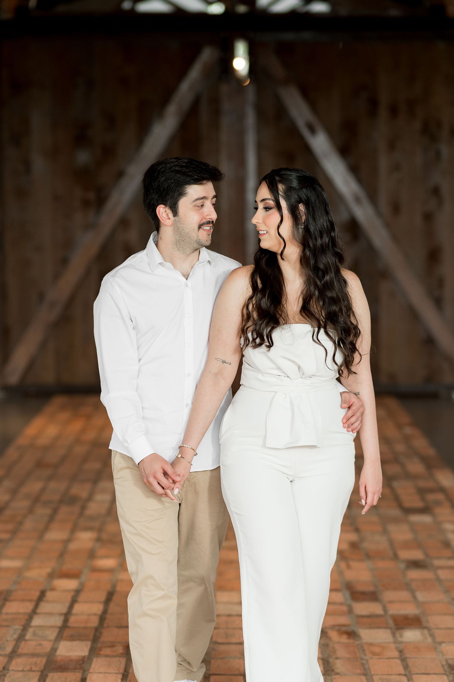A couple in white outfits holds hands and gazes at each other inside a rustic barn with brick floors.