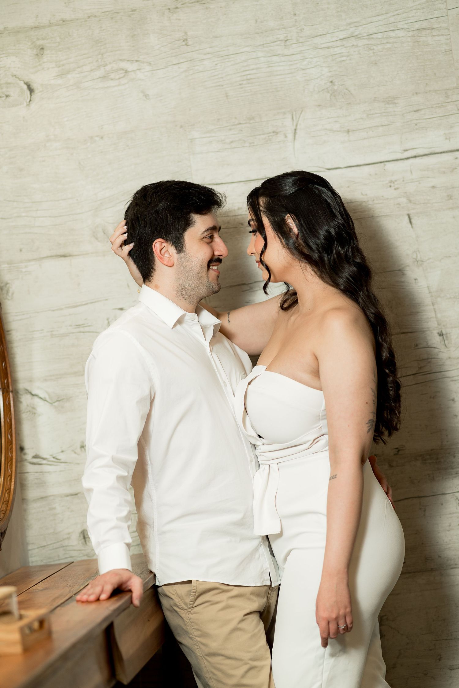 Couple in white outfits sharing a romantic moment, leaning against a rustic wooden wall indoors.