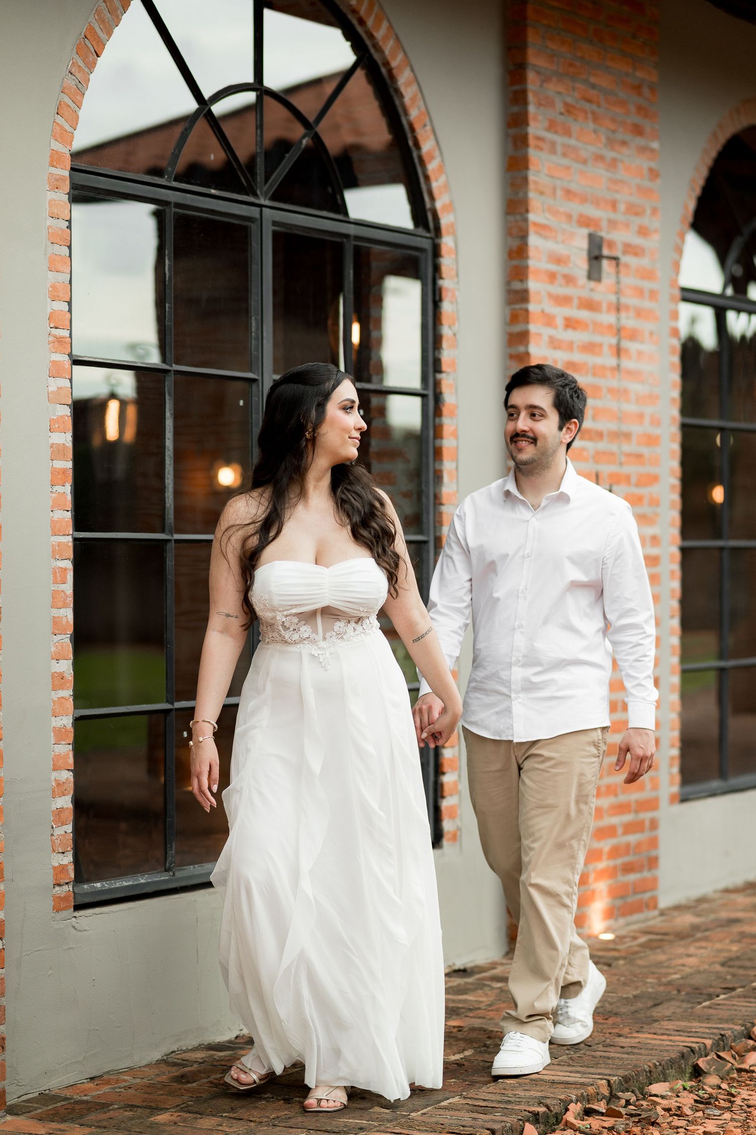Couple in wedding attire walking hand in hand outside a brick building with arched windows.