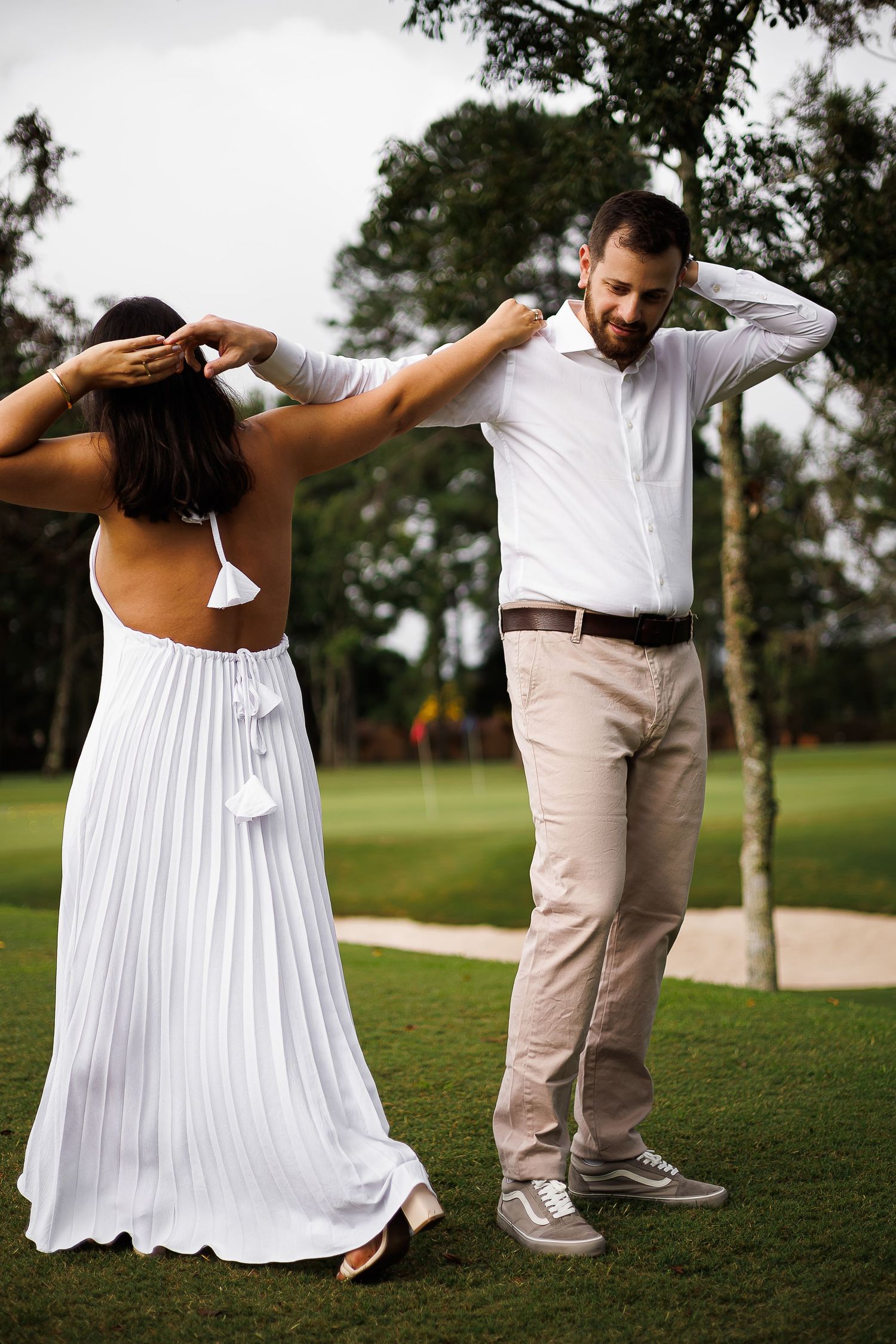 Couple in white outfits adjusting hair outdoors on a golf course green, woman in backless dress.