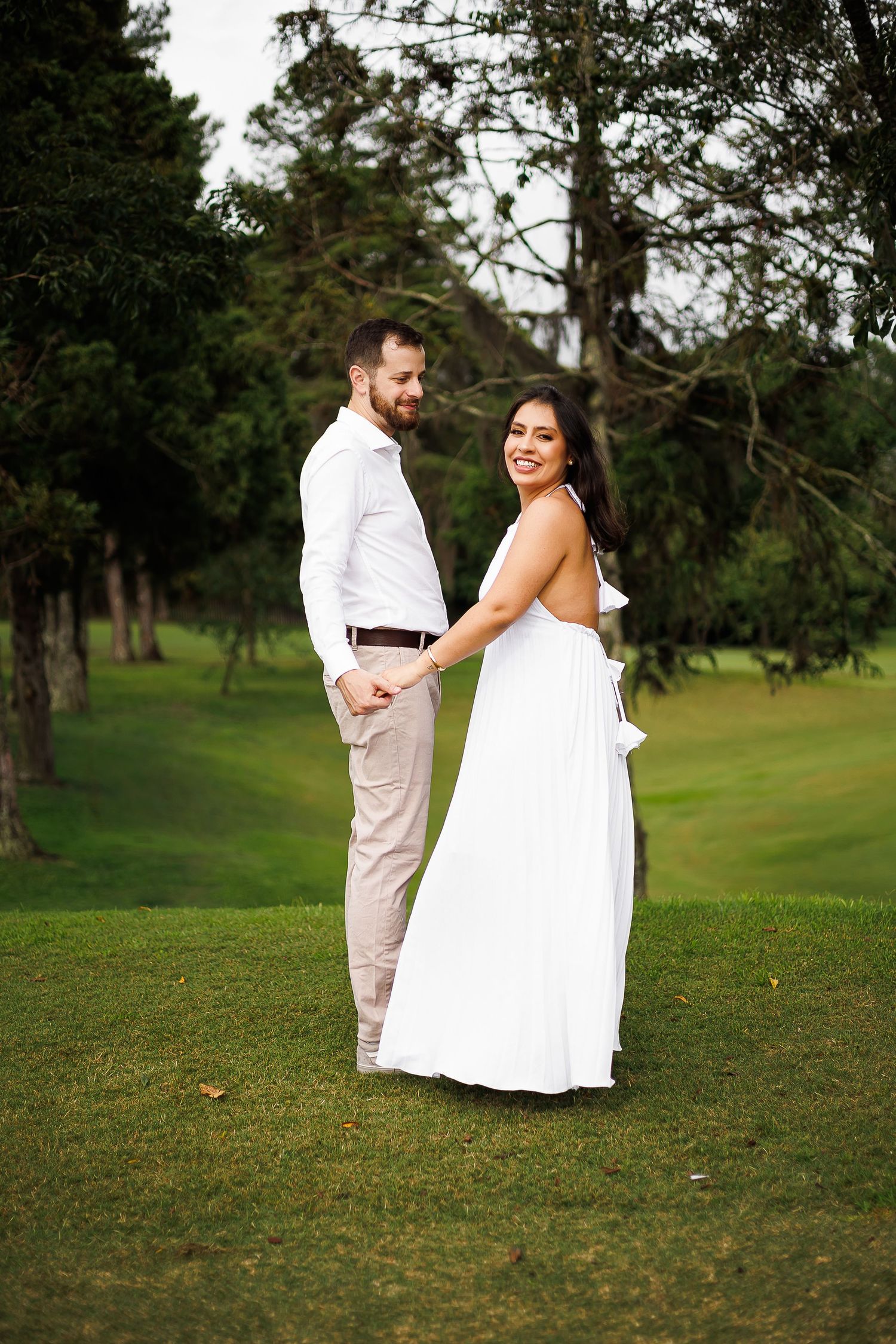 Couple holding hands on green lawn, woman in white backless gown smiling, man in white shirt and khaki pants, trees behind.