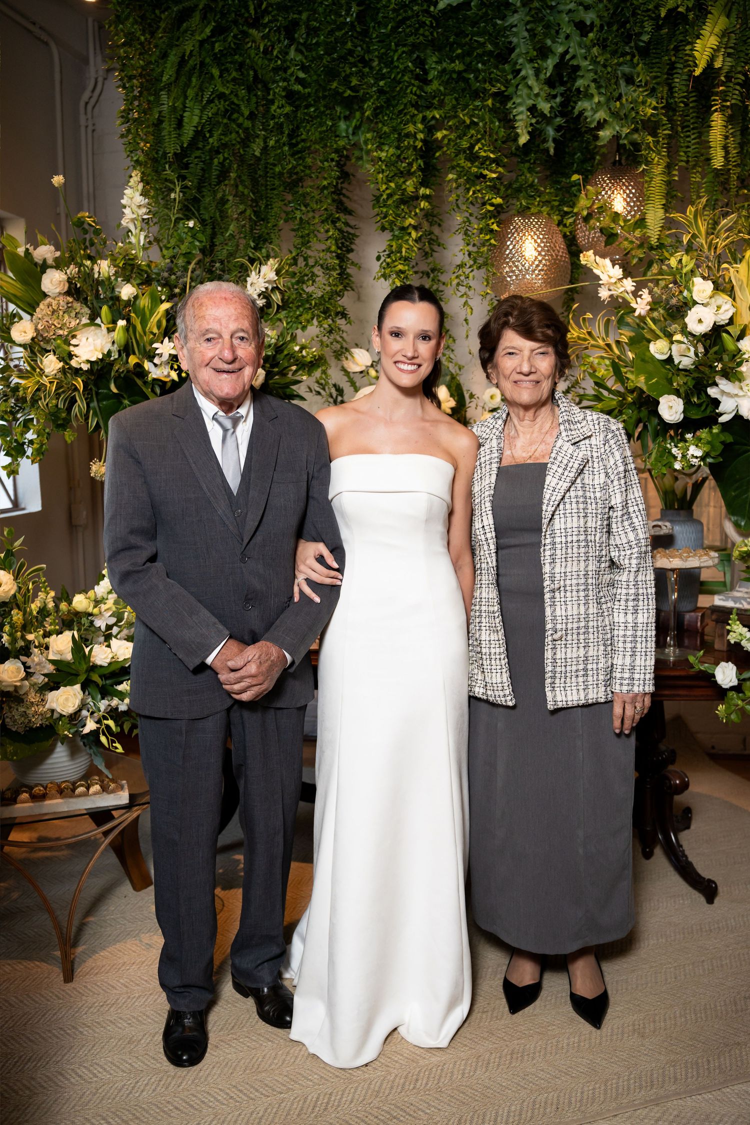 Bride in white strapless gown poses with elderly couple amid lush greenery and white floral arrangements at wedding.