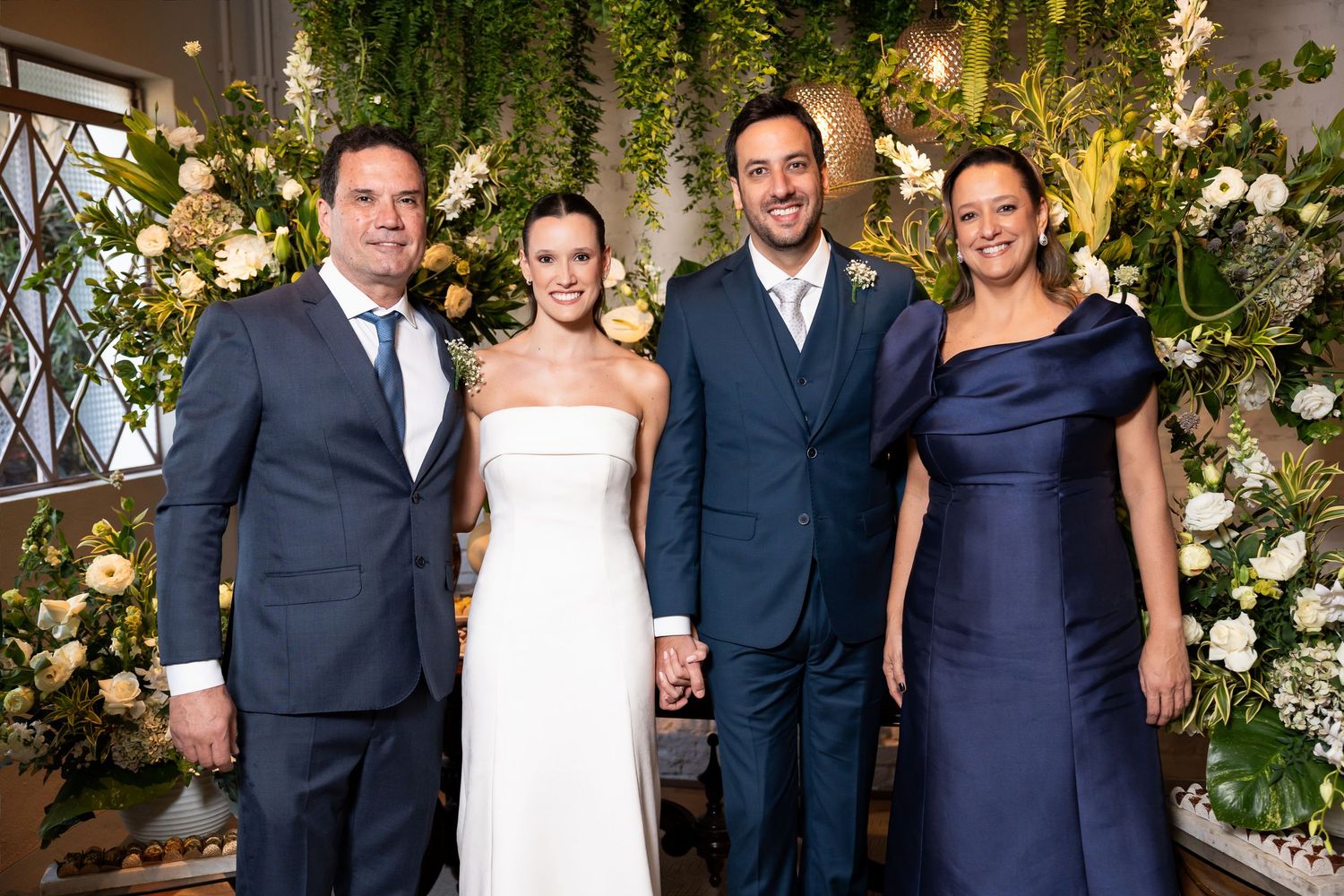 Wedding party of four posing in front of lush floral backdrop, bride in white gown holding hands with groom in gray suit.