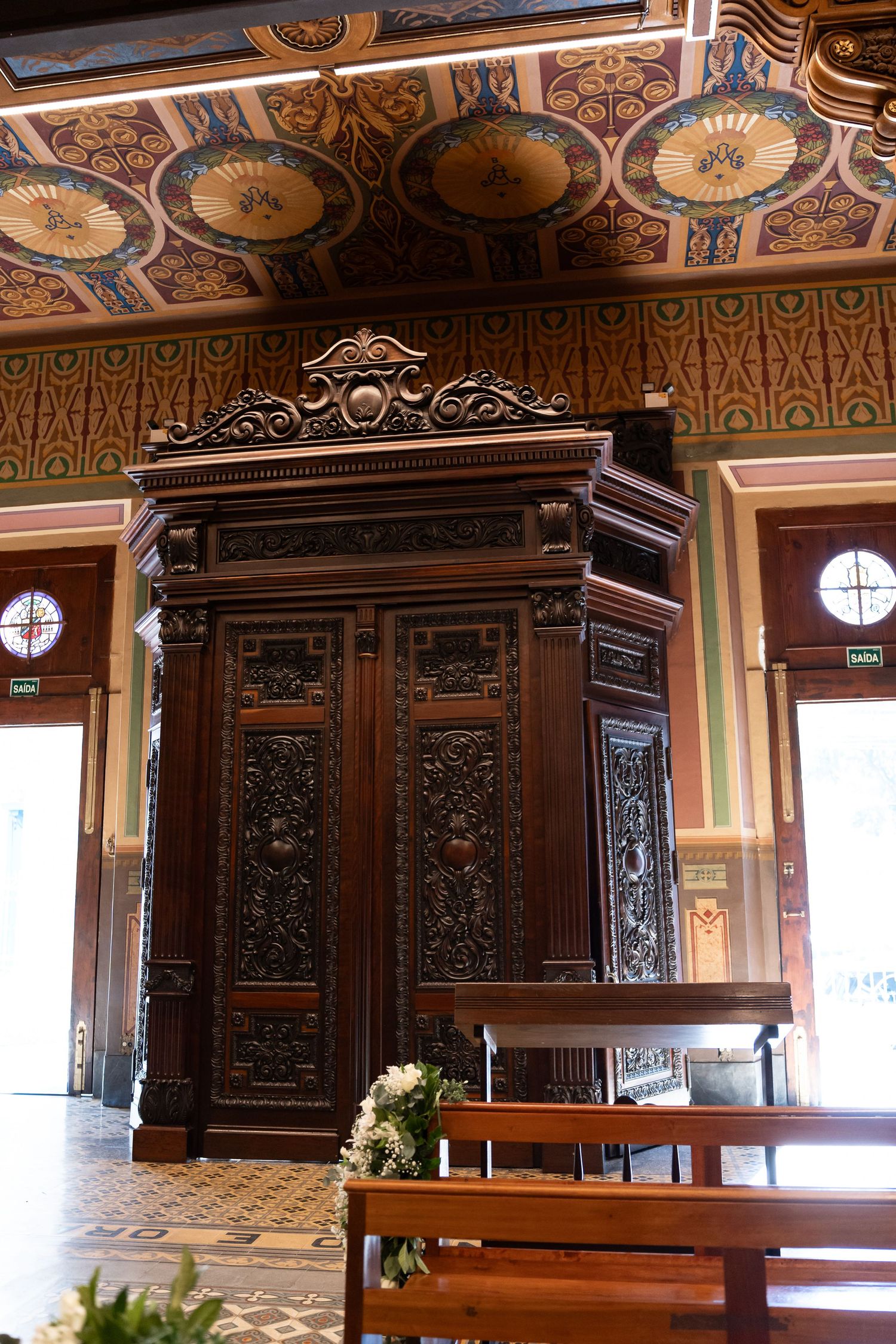 Ornate carved wooden confessional booth inside a historic church with decorative painted ceiling and wooden pews.