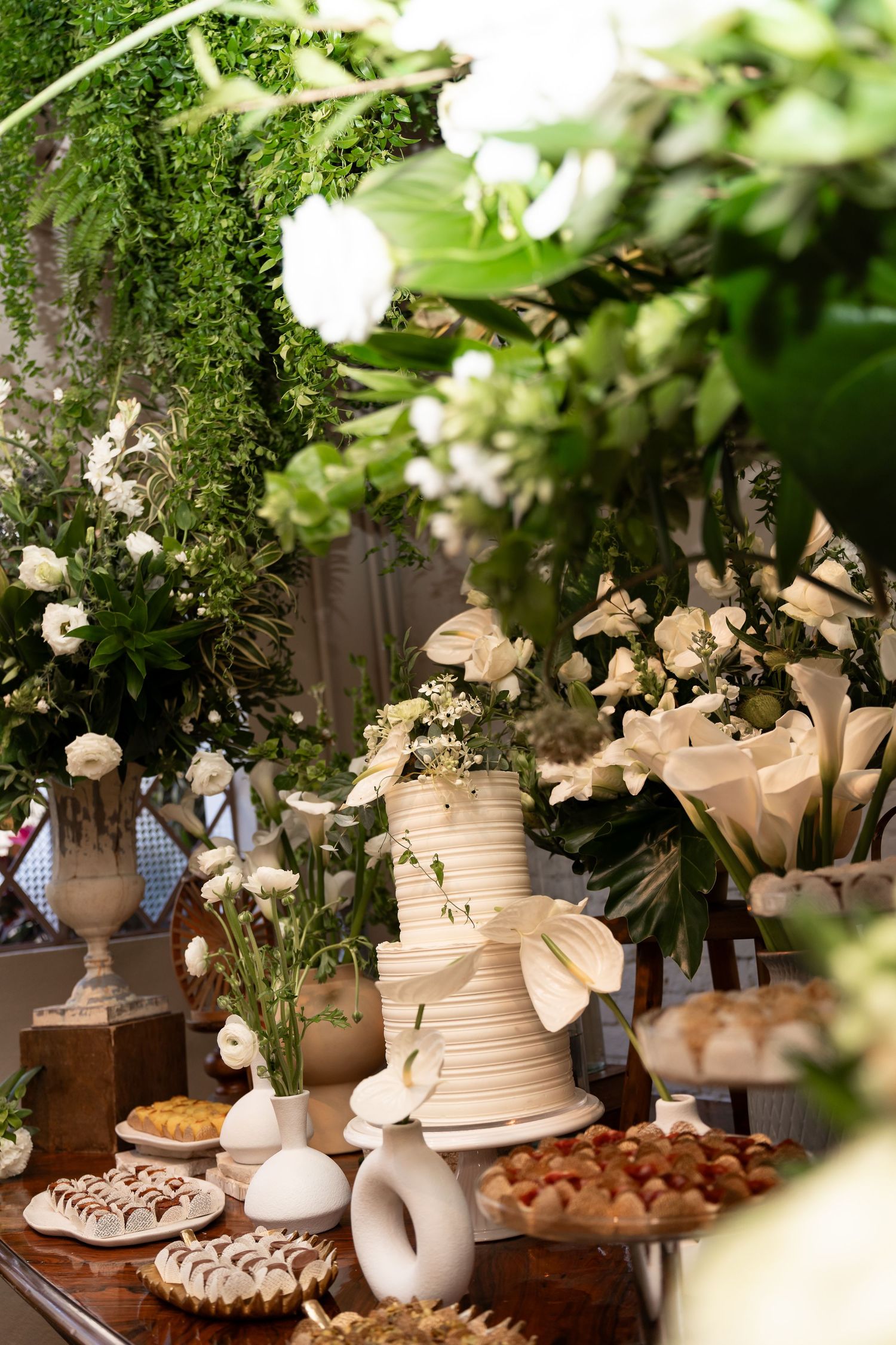 Elegant white wedding cake on dessert table with lush greenery, white flowers, and assorted pastries.