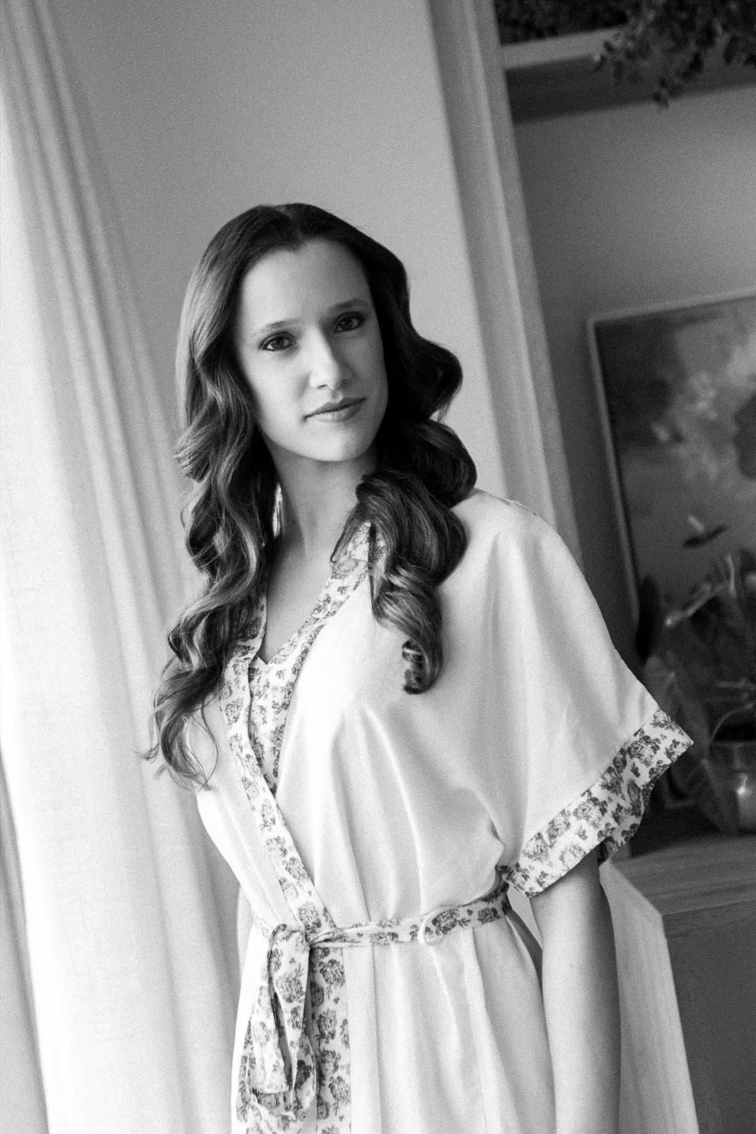 Black and white portrait of a woman in a floral-trimmed robe with long wavy hair, posing elegantly indoors.