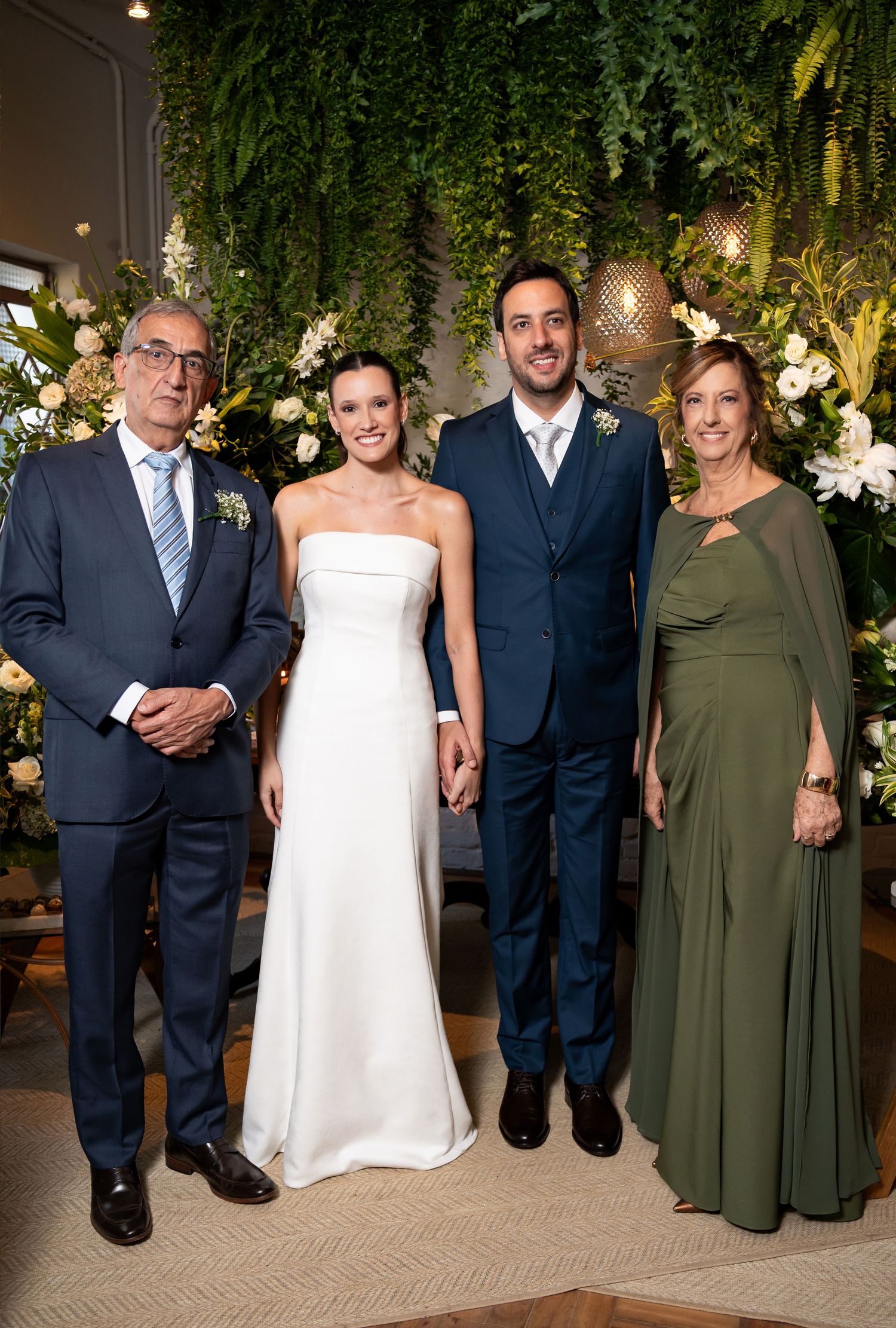Bride and groom pose with family members at elegant wedding ceremony with lush green floral backdrop.