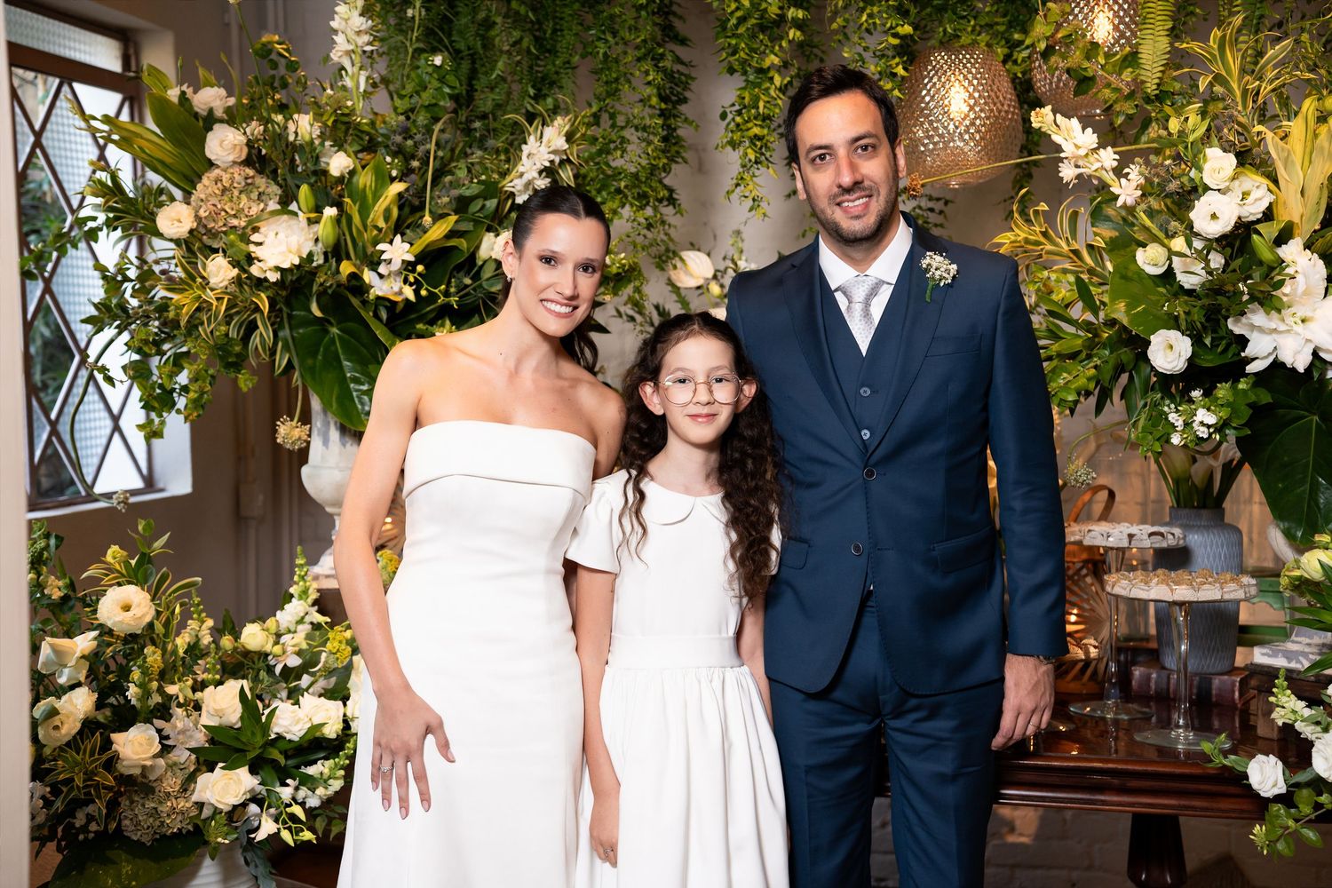 Bride and groom in wedding attire pose with young girl in white dress before floral backdrop.