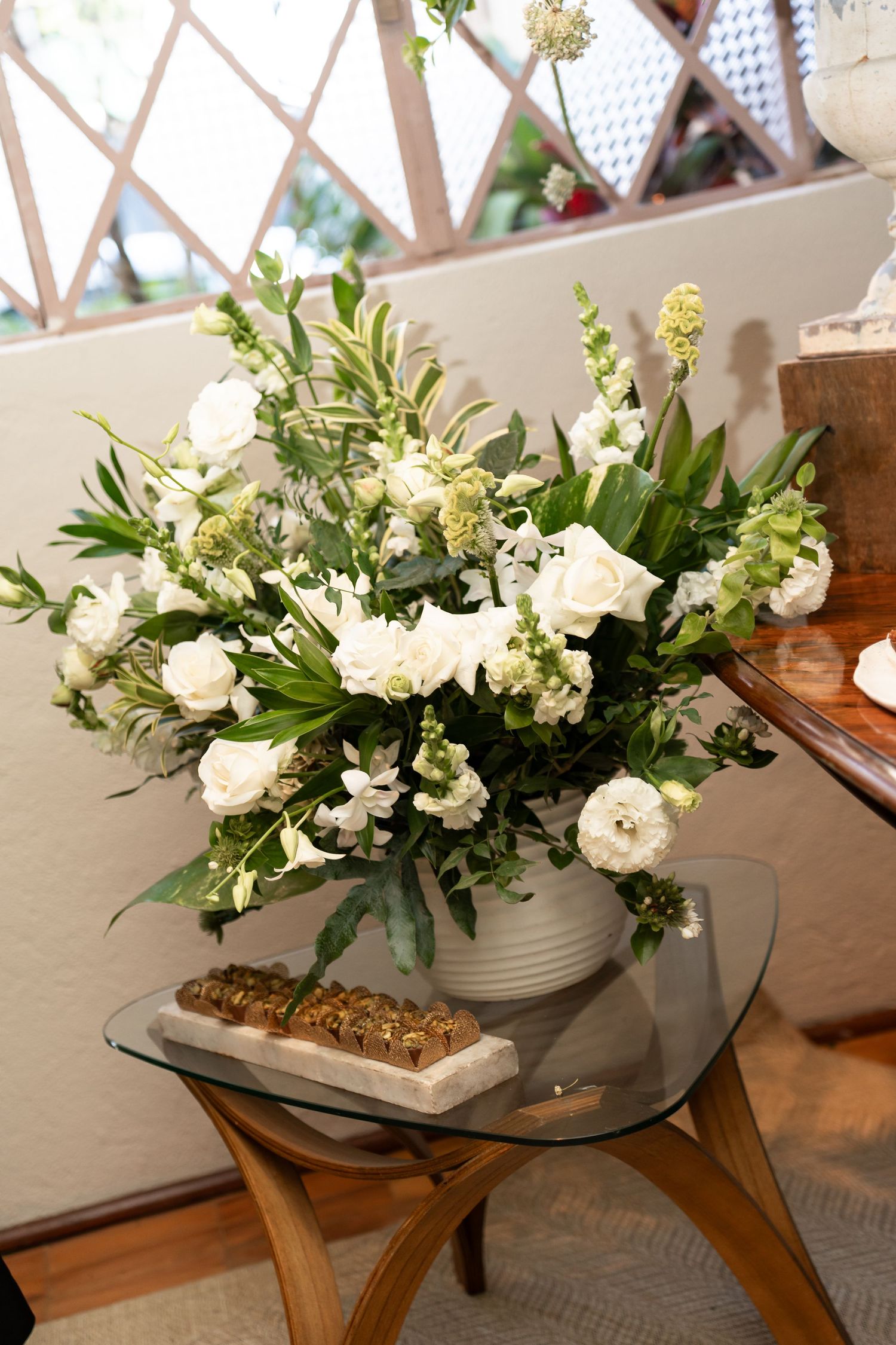 White floral arrangement with roses and greenery in a white vase on a glass table indoors.