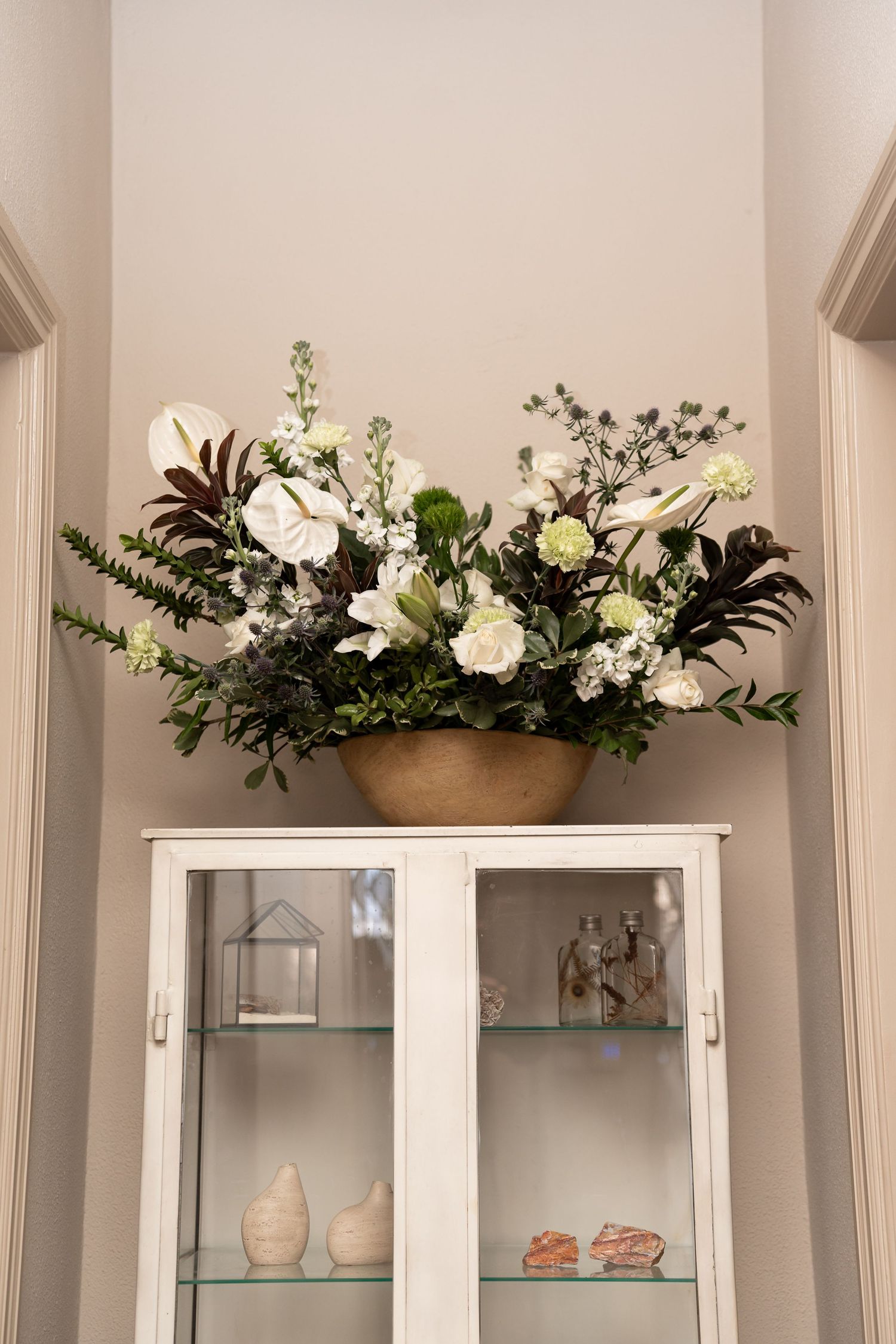 White floral arrangement in gold bowl atop glass display cabinet with decorative objects on shelves.
