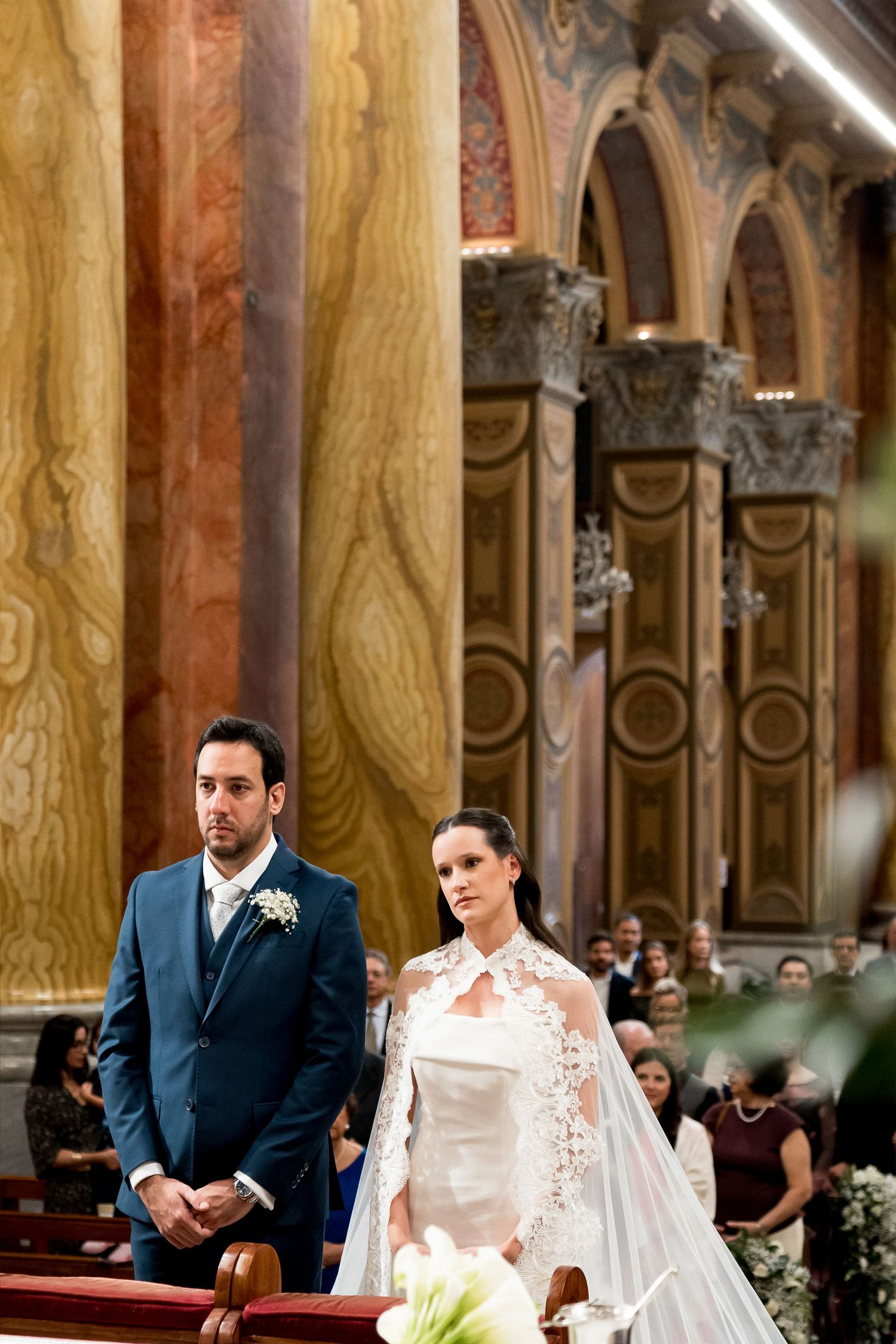Bride in lace wedding gown and groom in navy suit standing at church altar during elegant wedding ceremony.