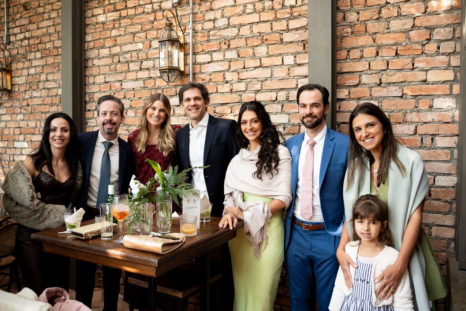 Group of elegantly dressed adults and a child posing together at a rustic brick-walled restaurant celebration.