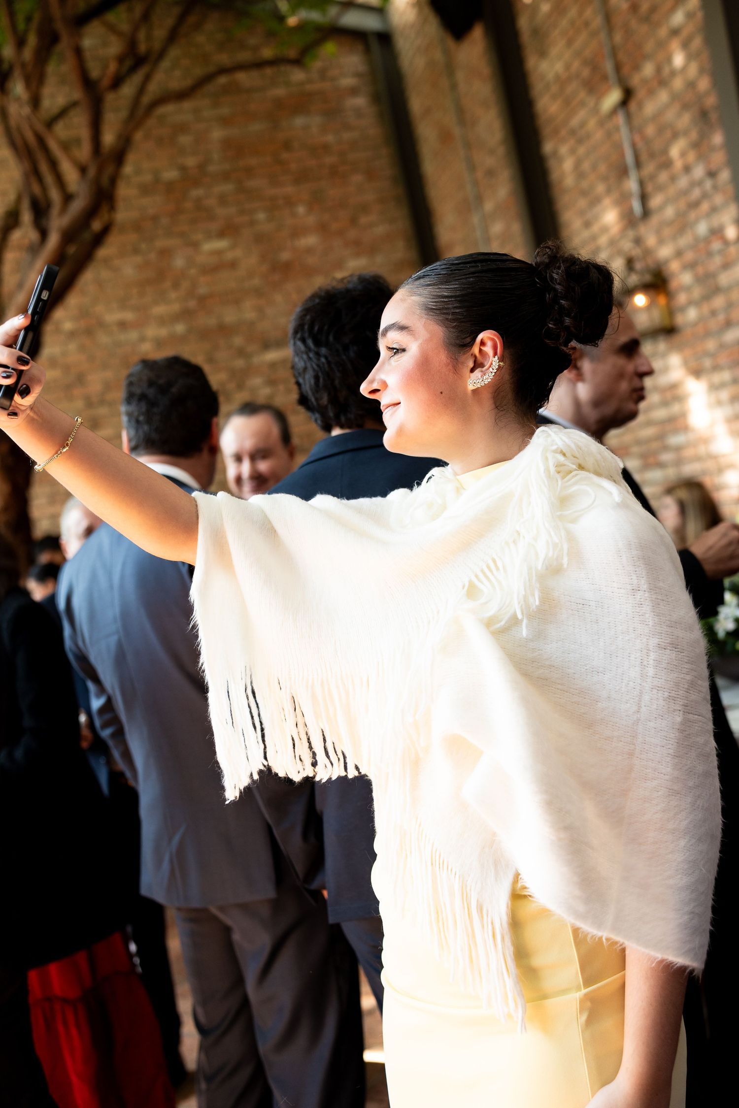 Woman in white fringed shawl taking a selfie at an elegant outdoor event near a brick wall.