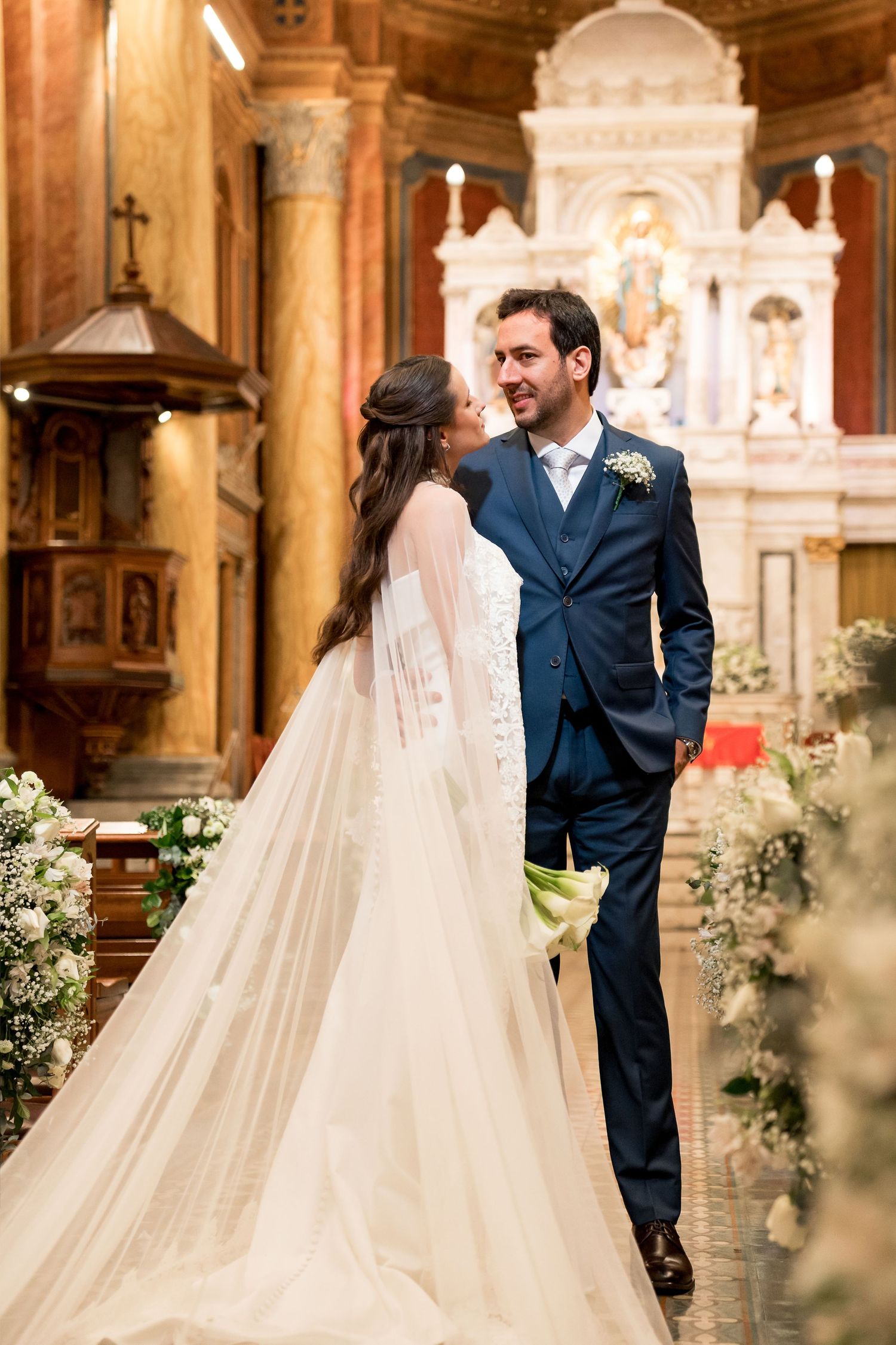 Bride and groom share a romantic moment at church altar, bride in white gown with veil, groom in navy suit.