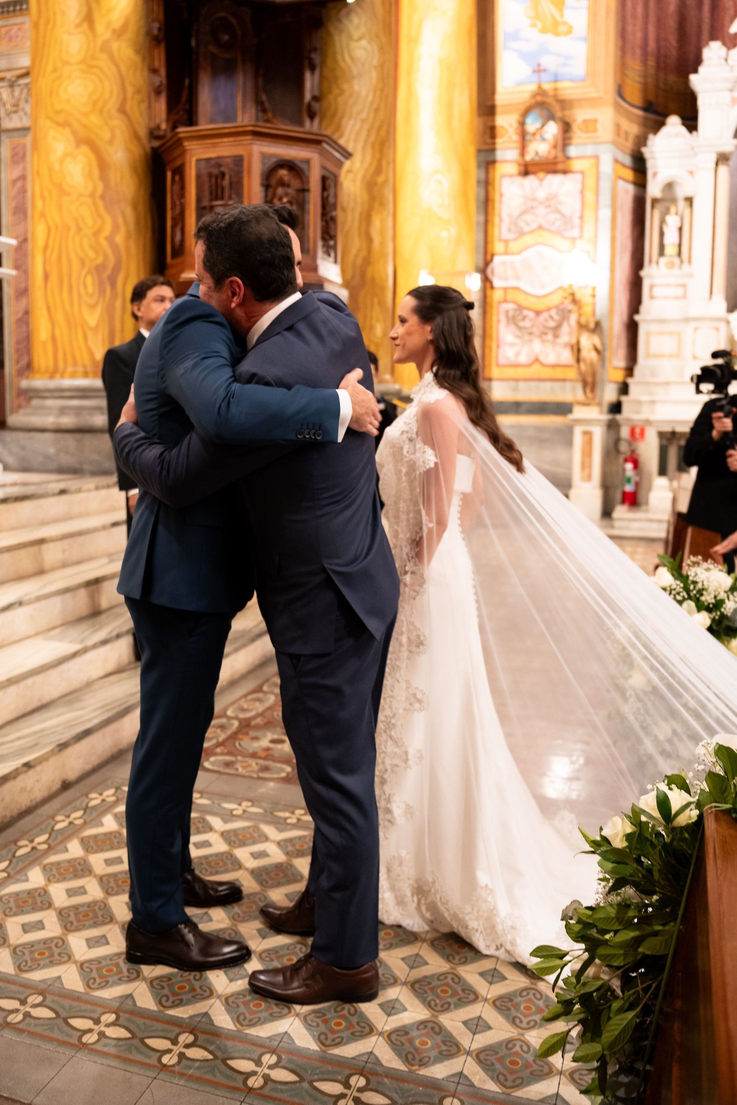 Bride in lace gown with veil embraces guest in blue suit at elegant church wedding ceremony.