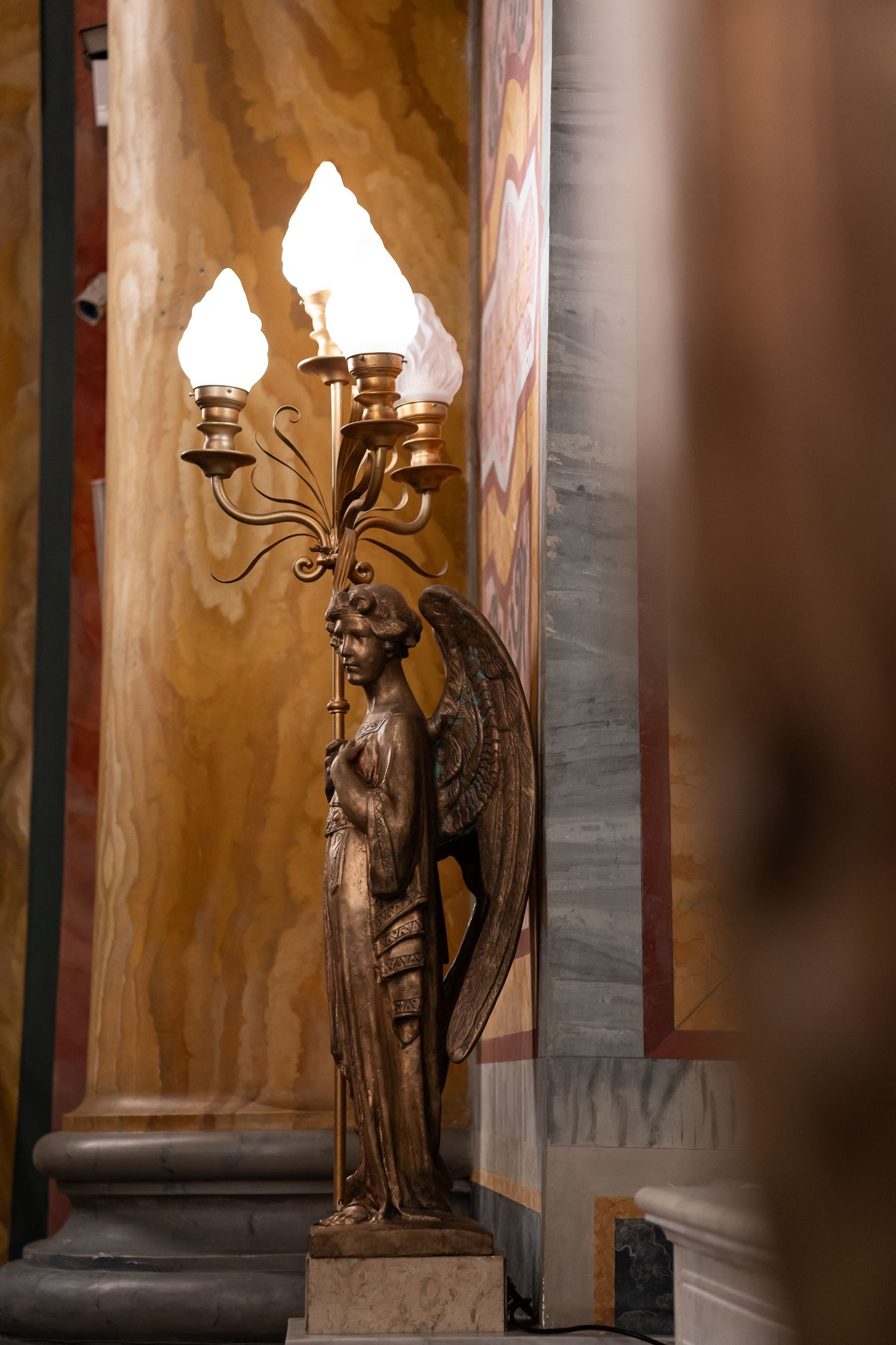 Golden angel candelabra sculpture with lit flame lights against ornate marble wall in elegant interior.