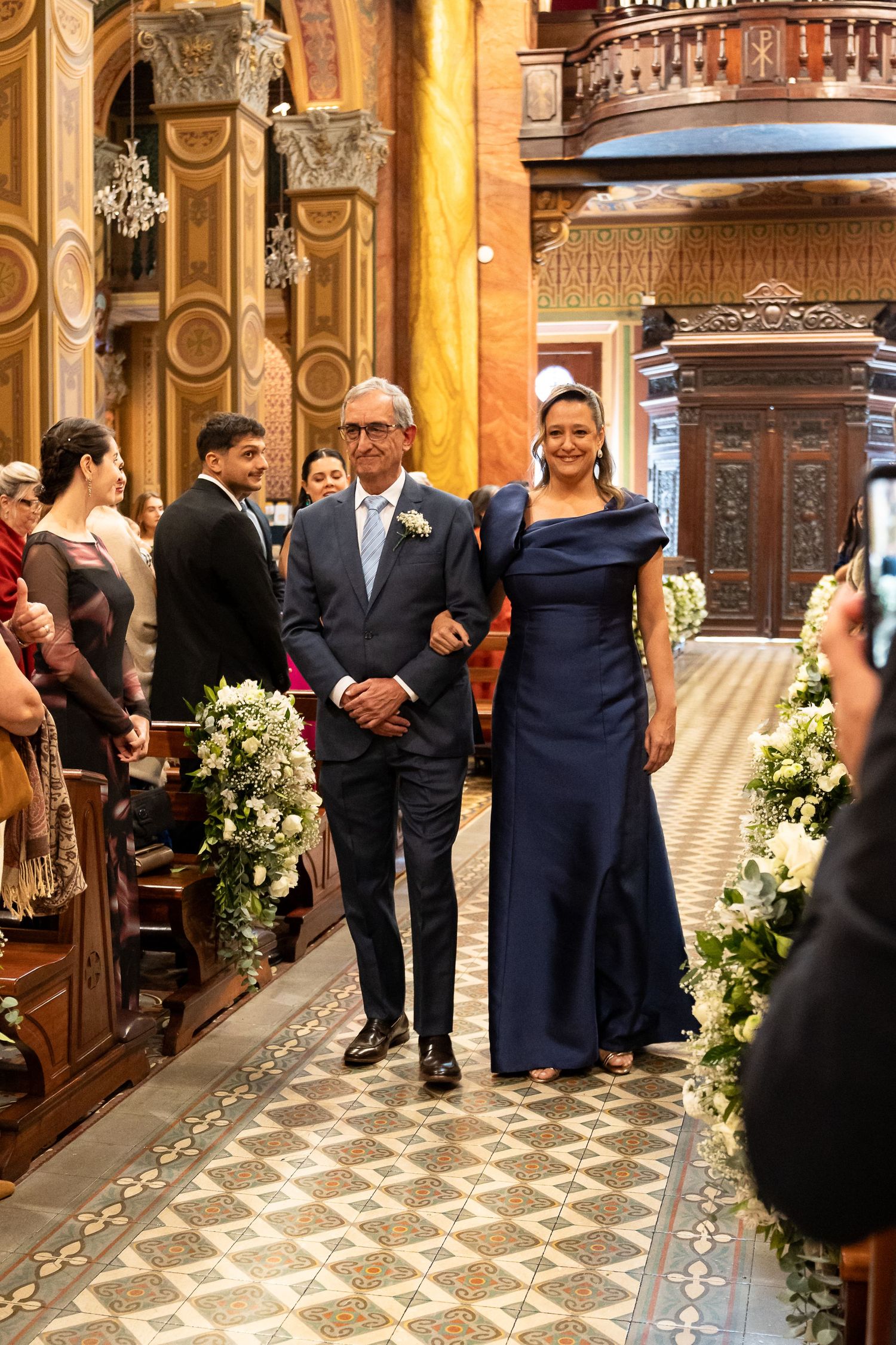 Elegant couple walking down church aisle during wedding ceremony, woman in navy gown, man in blue suit, floral decorations.