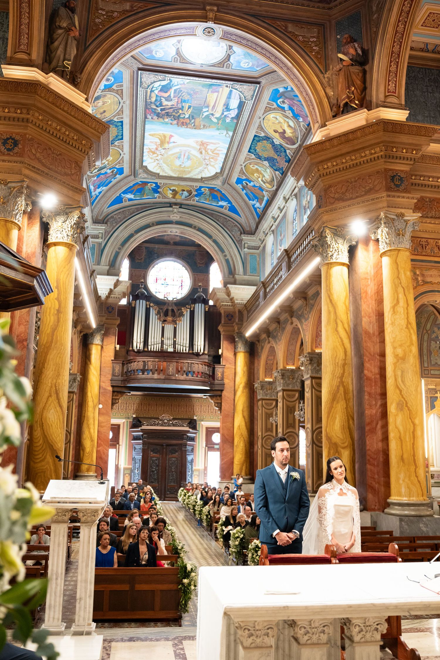 Couple stands at altar in ornate church with colorful frescoed ceiling, marble columns, and pipe organ during wedding ceremony.