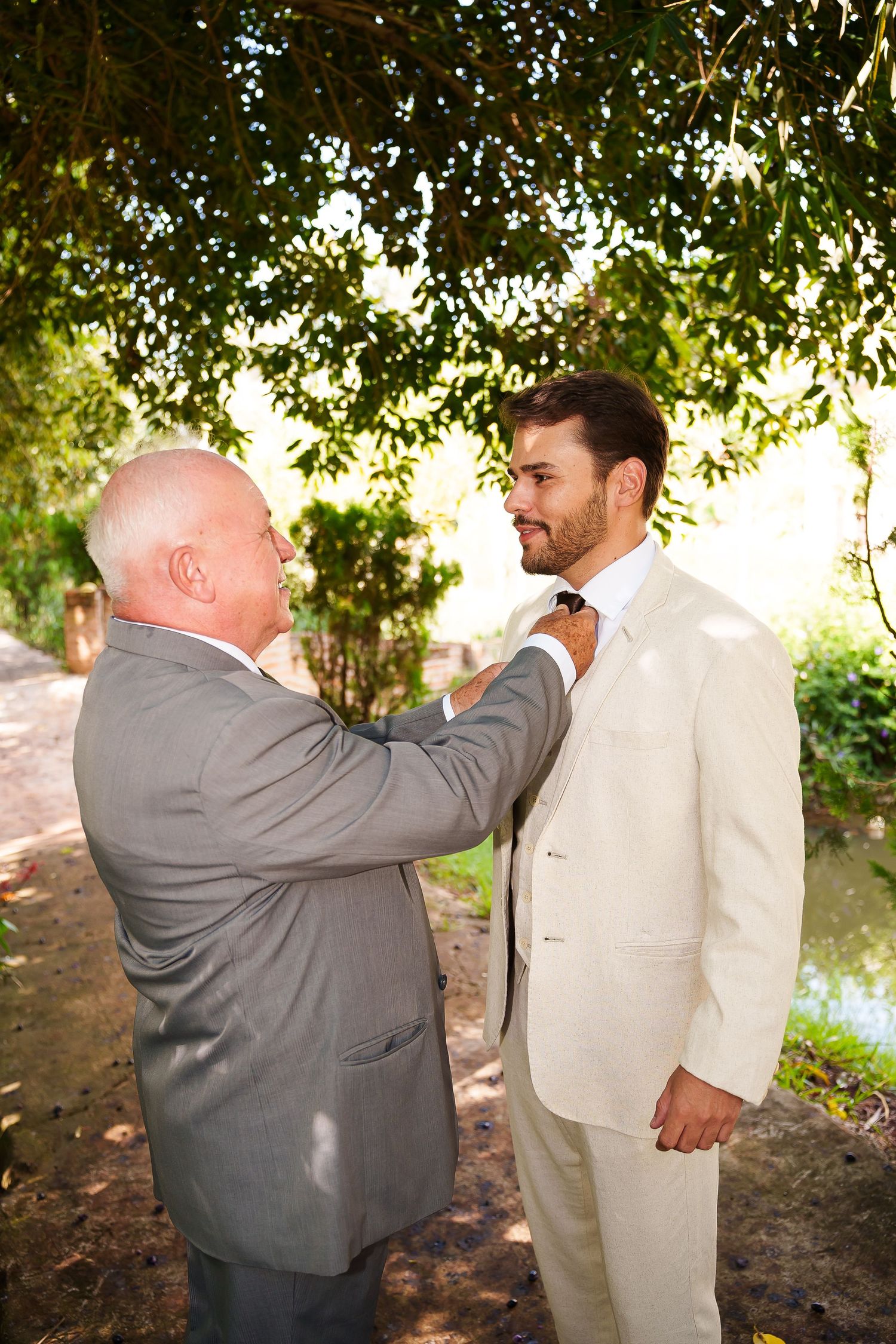 Older man in gray suit adjusting tie of young groom in white suit outdoors before wedding ceremony.